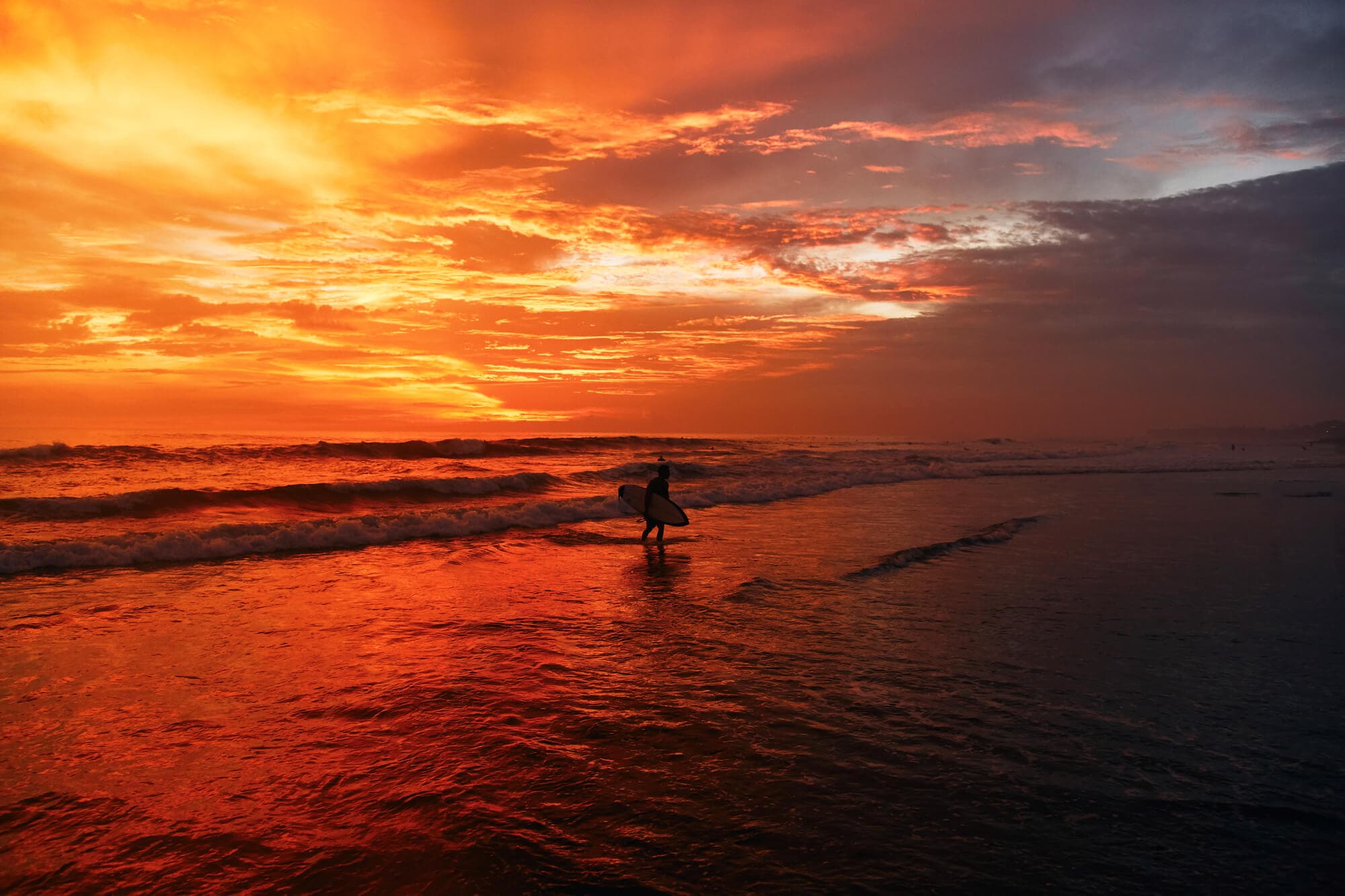 Silhouette of a surfer carrying his board on Batu Bolong Beach in Canggu during a yellow, orange and red sunset.