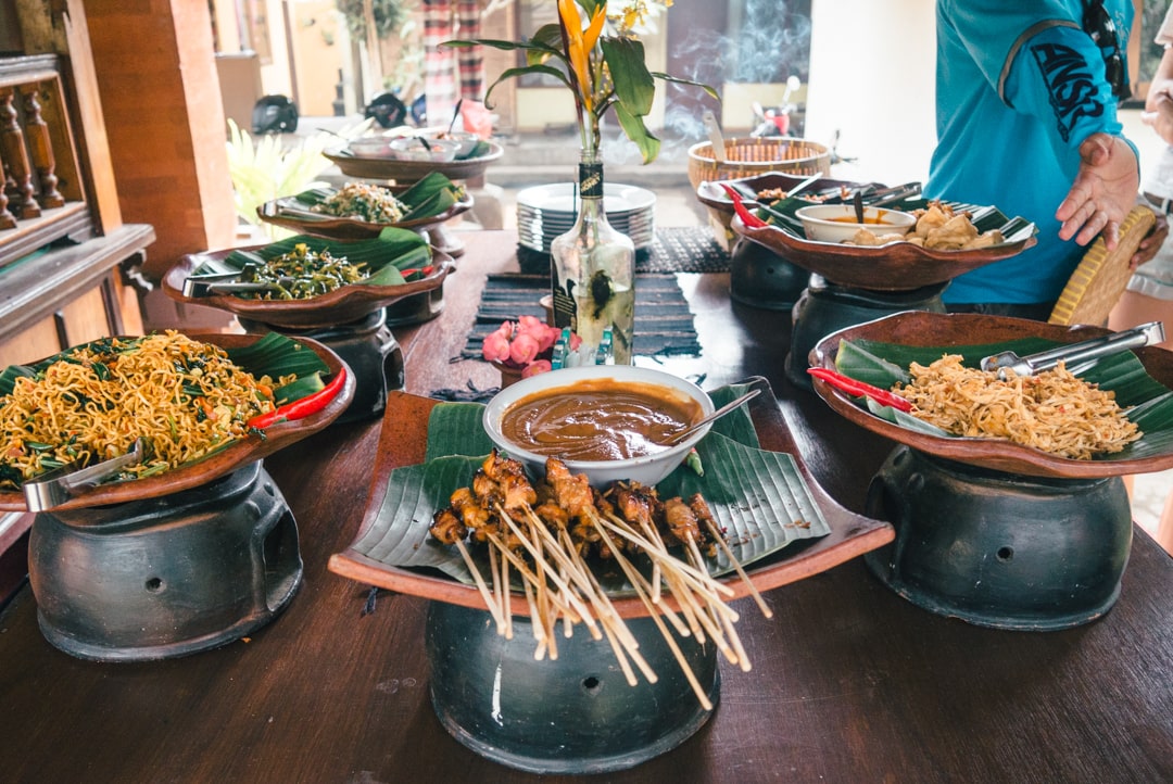 A large wooden table in the middle of a traditional Balinese compound, with a feast of Indonesian dishes like sate chicken and fried noodles.