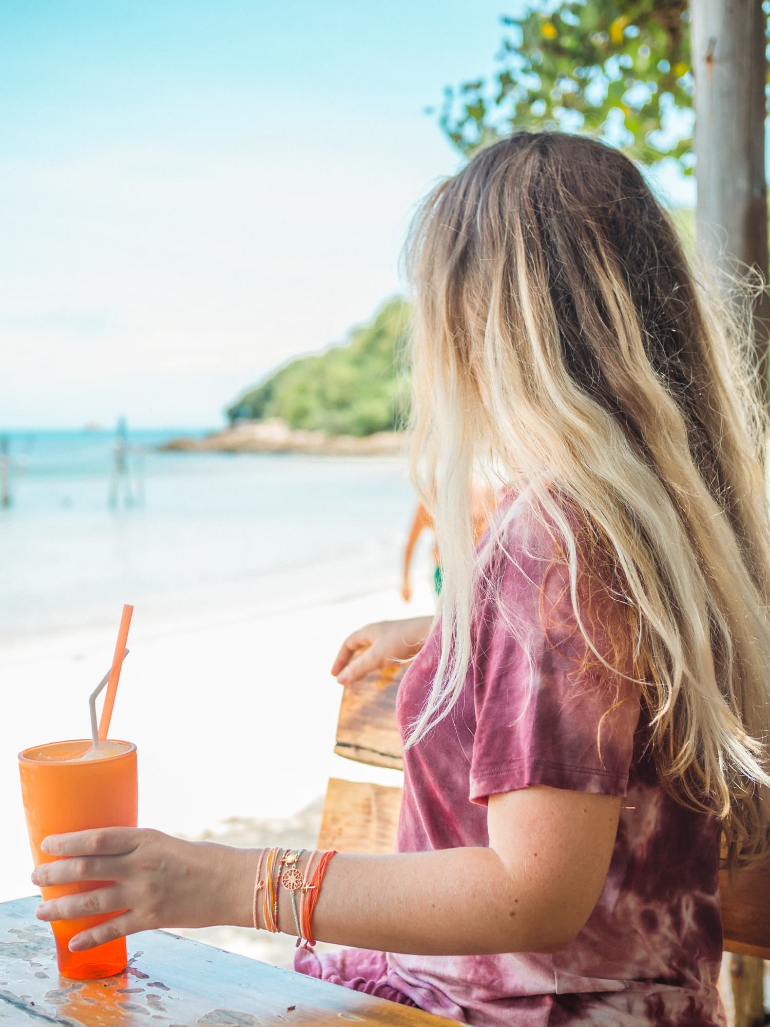 Girl with long dark blonde hair looking out at sea with an orange granita in one hand at Jelly Fish Beach Restaurant on Koh Samet.