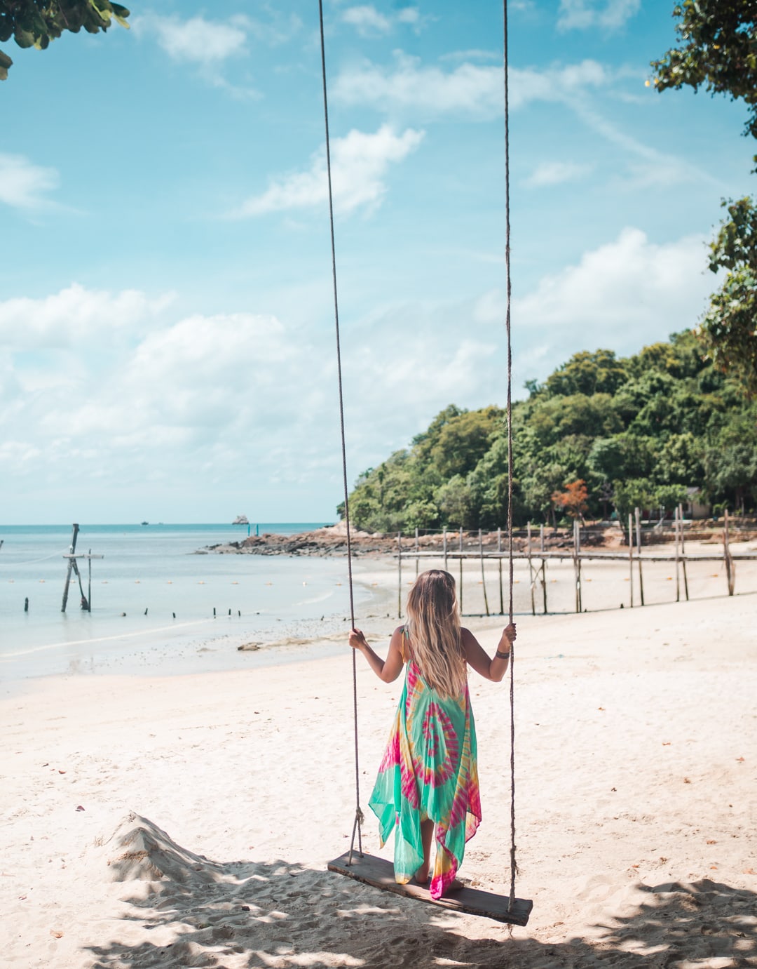 Girl with long hair wearing a green, yellow and pink dress standing on a swing on white-sand Ao Lung Dum Beach Koh Samet, Thailand.