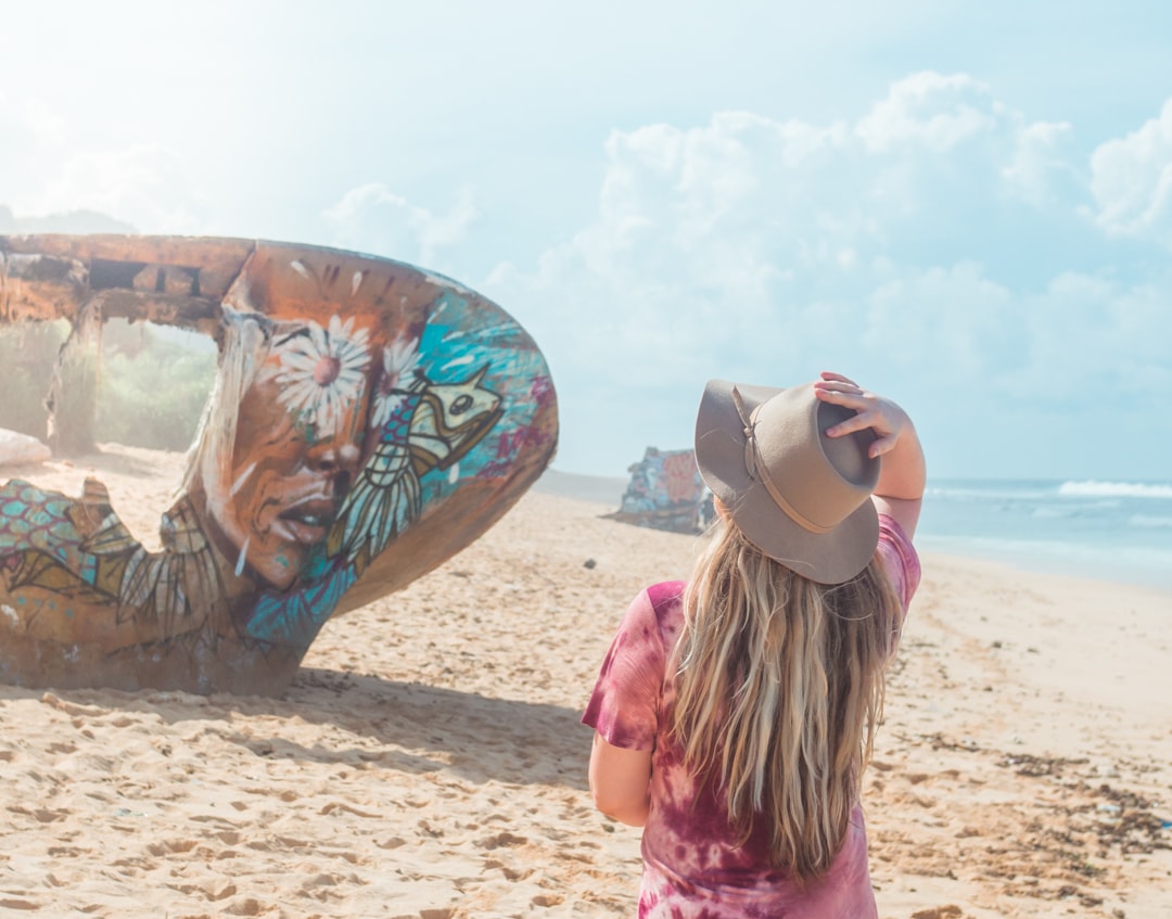 Woman holding on to a beige hat, wearing a purple tie dye dress, walking towards a shipwreck covered in graffiti on Nunggalan Beach, one of the top things to do in Bali.