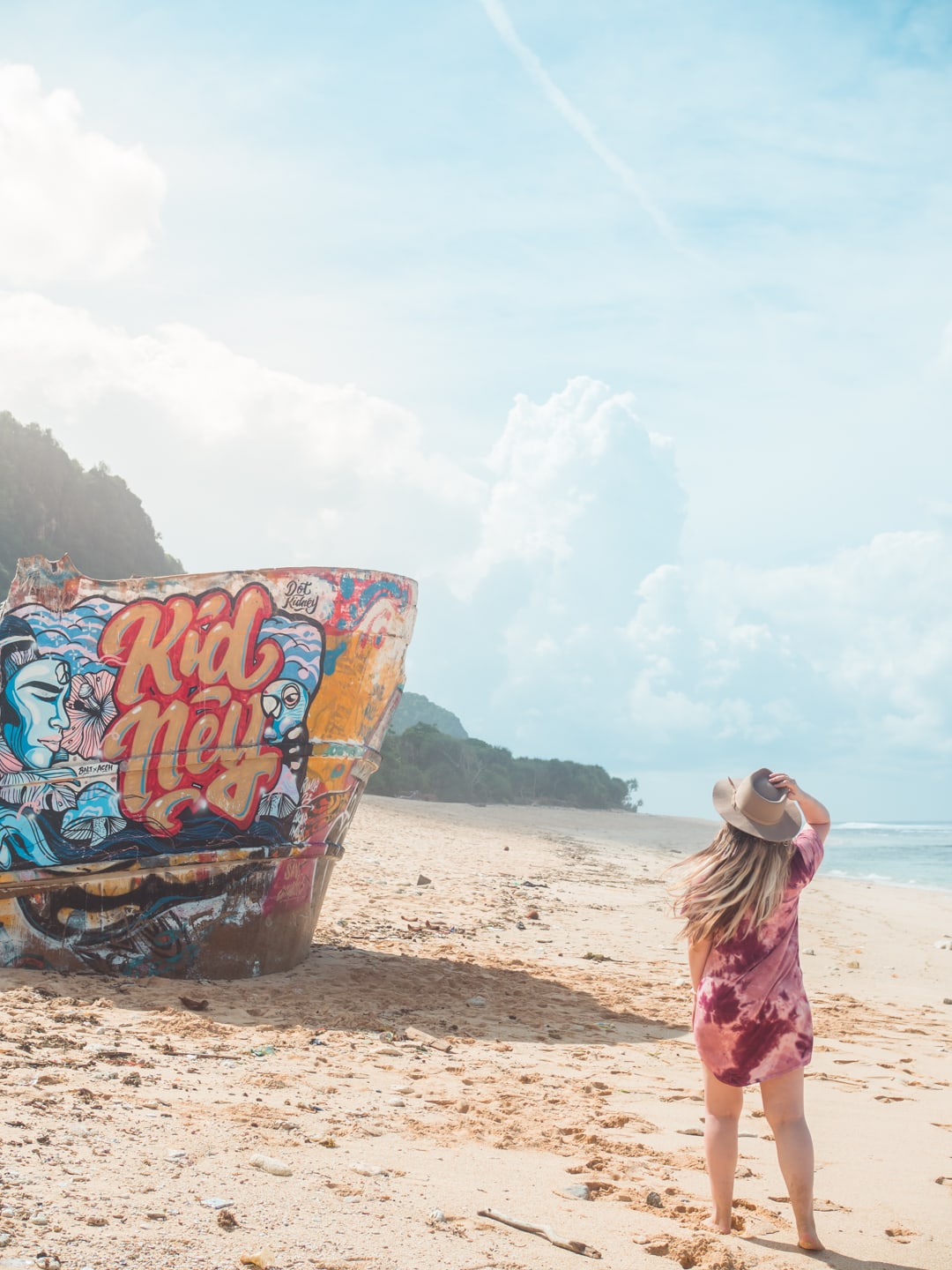 Woman with long hair, wearing a purple dress, holding on to her beige hat, walking towards a shipwreck with graffiti, one of the best hidden beaches in Uluwatu, Bali