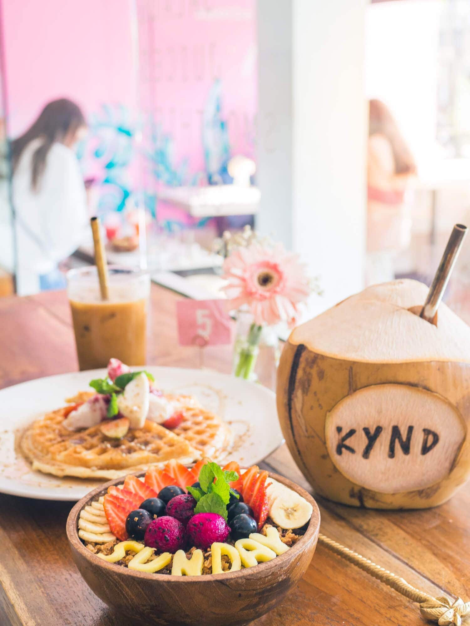 Smoothie bowl, coconut and waffles against a pink wall at Kynd Community Café in Seminyak, a must on any Bali Bucket List.