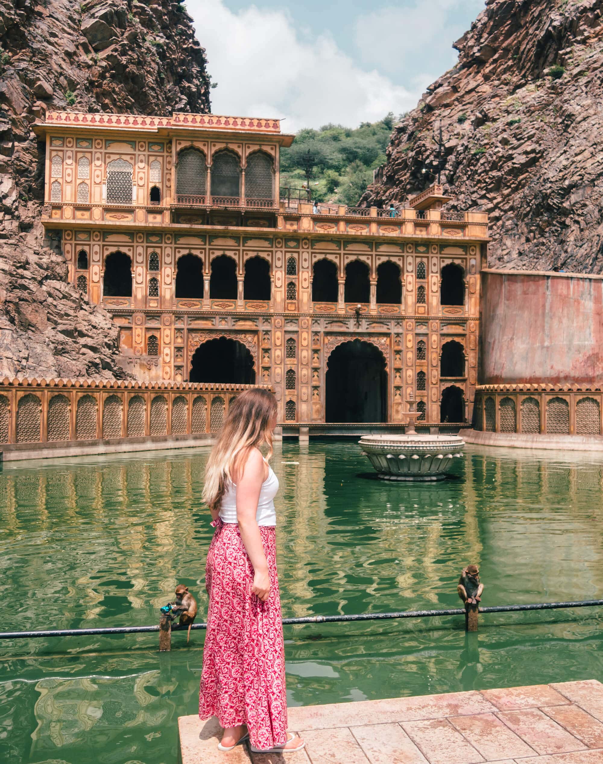 Girl with long hair, wearing a white top and pink maxi skirt, standing in front of a green pool at Galtaji Monkey Temple, one of the places you must see during your two days in Jaipur.