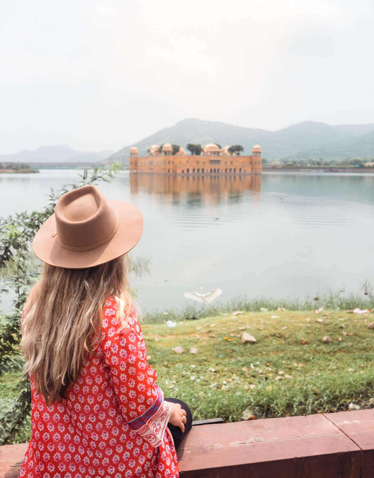 Girl with long hair wearing a beige felt hat, red and white kimono and black pants looking out over a Jal Mahal, a must on any 2-day Jaipur itinerary.