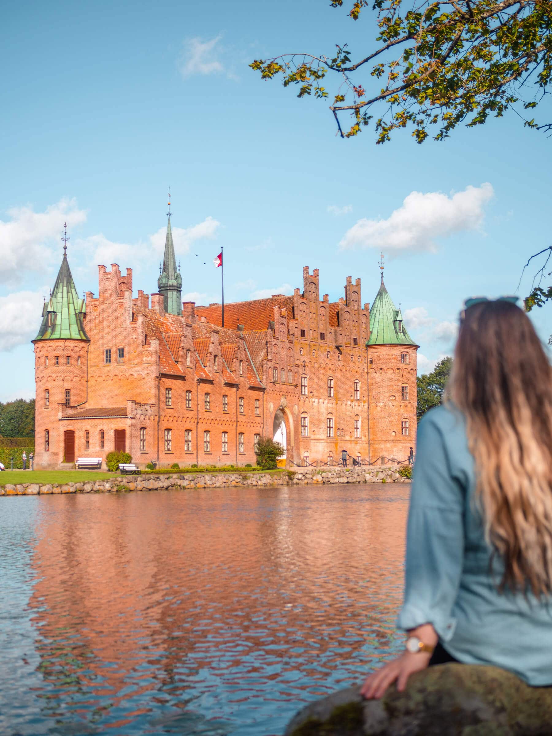 Girl with long dark blonde hair, wearing a denim short, sitting in front of the orange Egeskov Castle that are surrounded by a moat.