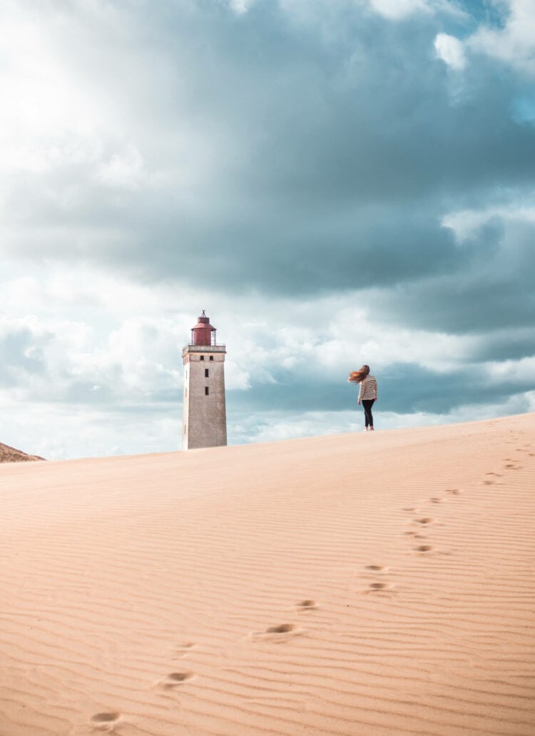 Girl with long hair blowing in the wind, wearing a white and blue striped swater and black pants, walking on a sand dune towards Rudbjerg Knude Lighthouse on a stormy day. The perfect day trip from Aarhus.