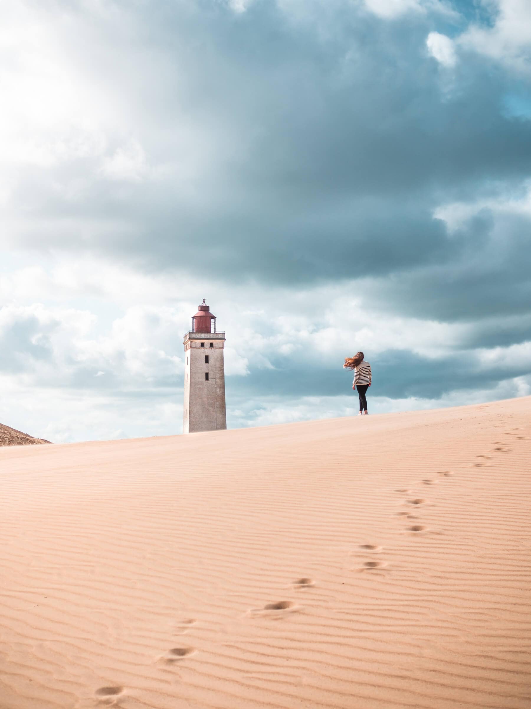 Girl with long hair blowing in the wind, wearing a white and blue striped swater and black pants, walking on a sand dune towards Rudbjerg Knude Lighthouse on a stormy day. The perfect day trip from Aarhus.
