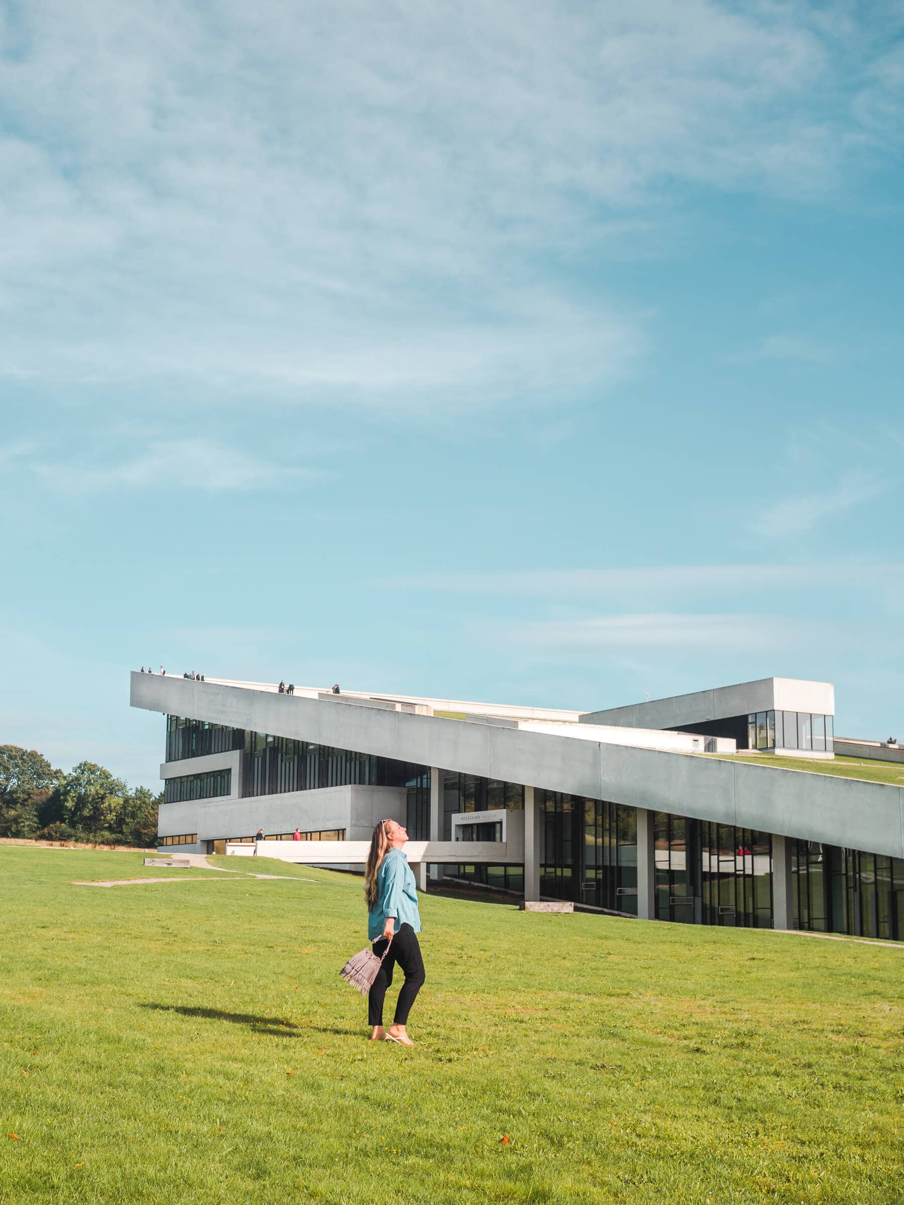 Girl with long hair, wearing a denim shirt and black pant holding a grey bag, walking in front of the modern Moesgaard Museum on a sunny day. The perfect day trip from Aarhus.