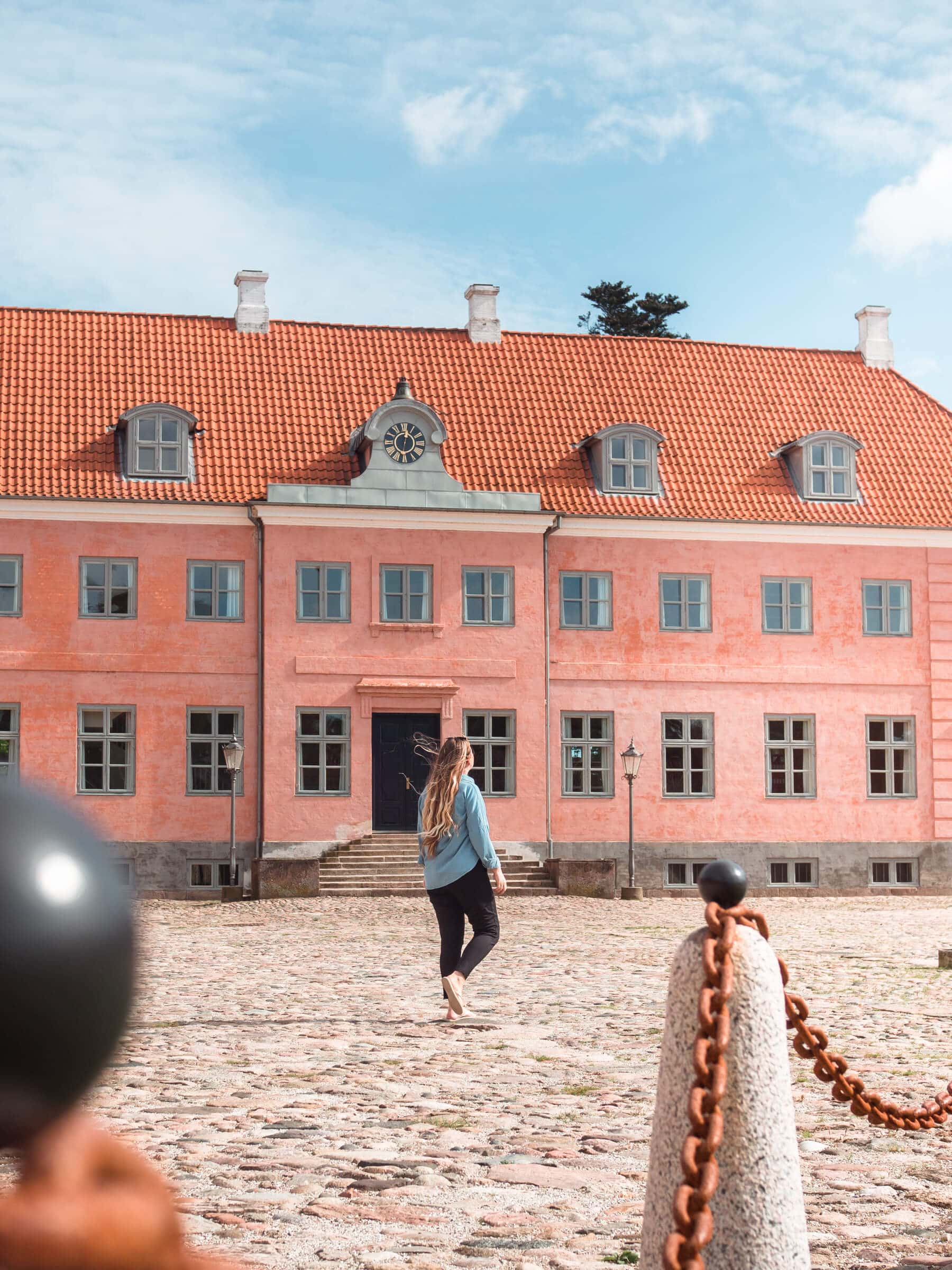 Girl with long hair wearing black pants and a denim shirt walking in front of the beautiful pink Moesgaard Manor, on a day trip from Aarhus.