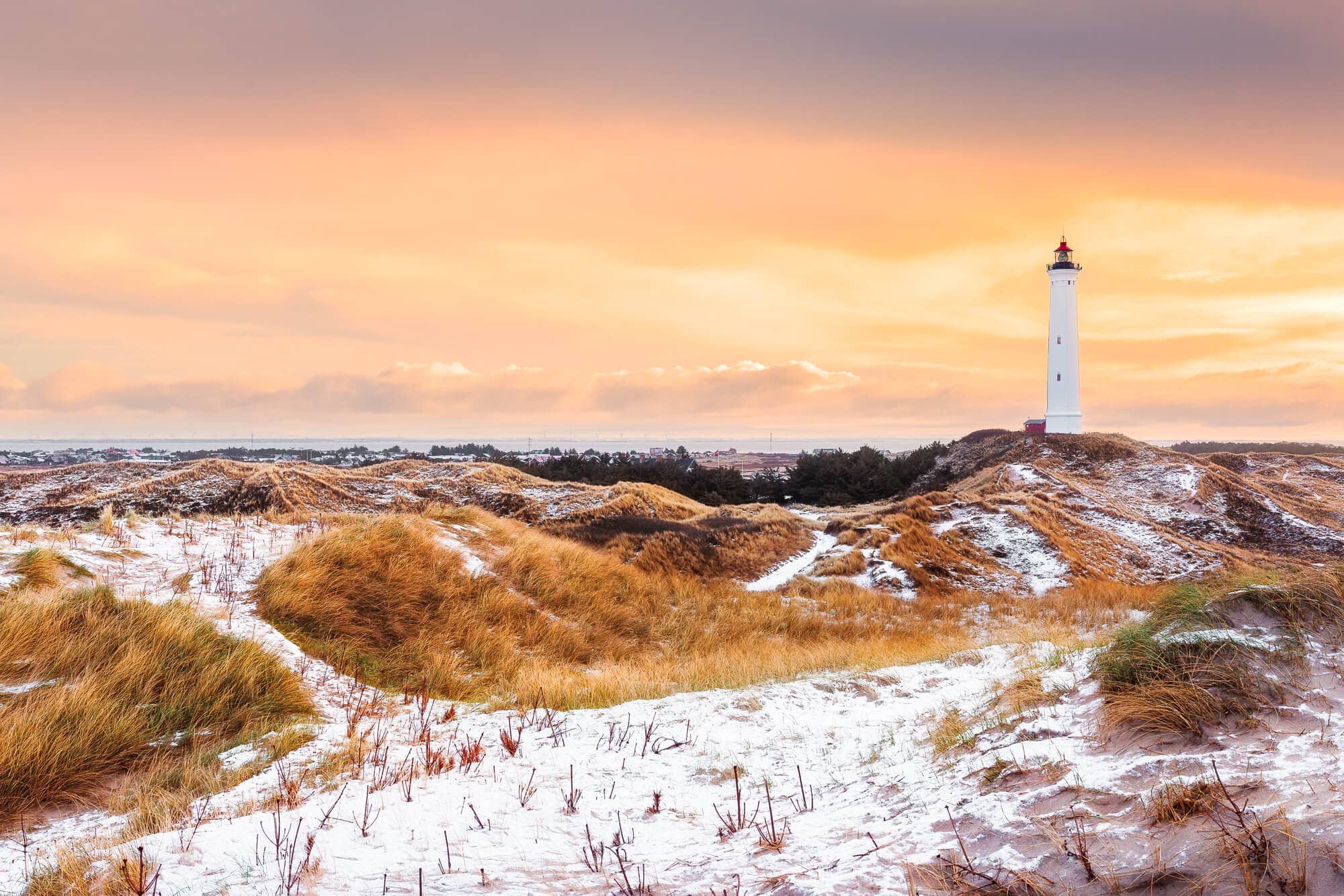 Lyngvig Lighthouse, close to Søndervig, set against an orange sky during sunset and surrounded by sand dunes covered in grass and snow. The perfect day trip from Aarhus.