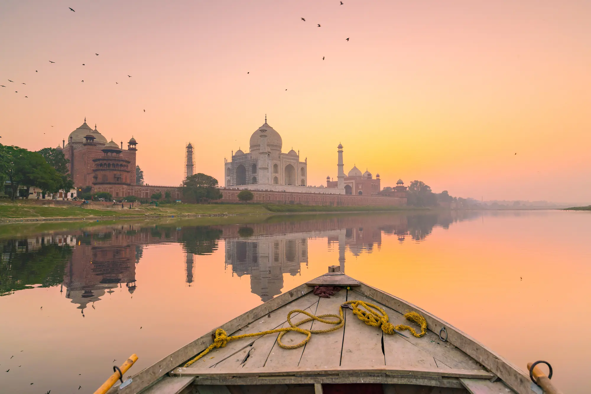 Photo of the Taj Mahal during a pink and yellow sunset shot from an old wooden boat on the calm river.