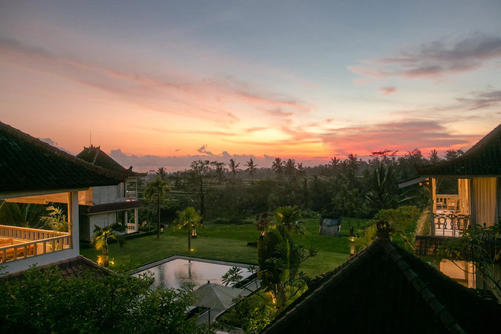 View above the roof at B.Saya Budget Hotel in Ubud Bali of a pool in a green garden with a pink and orange sunset over the rice fields.