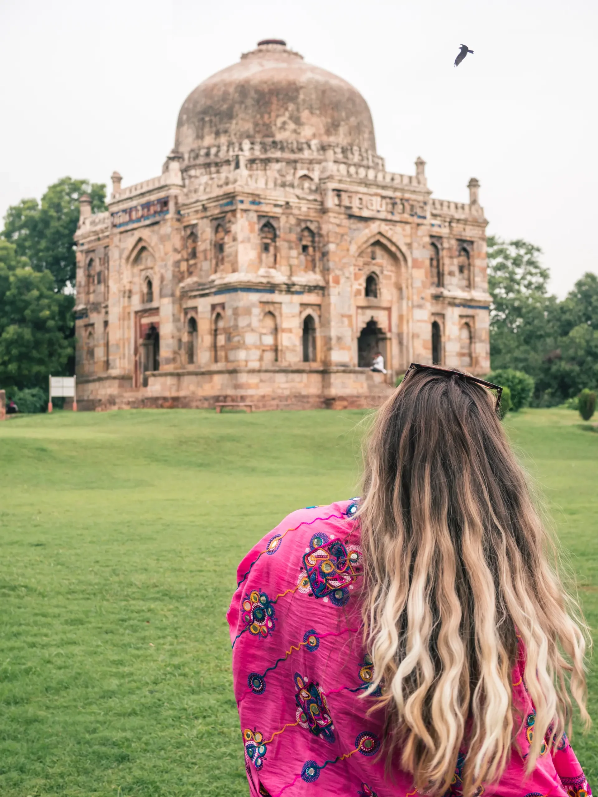 Woman with long dark blond hair, wearing a pink scarf, looking towards a beige building in Lodhi Gardens during first time in Delhi