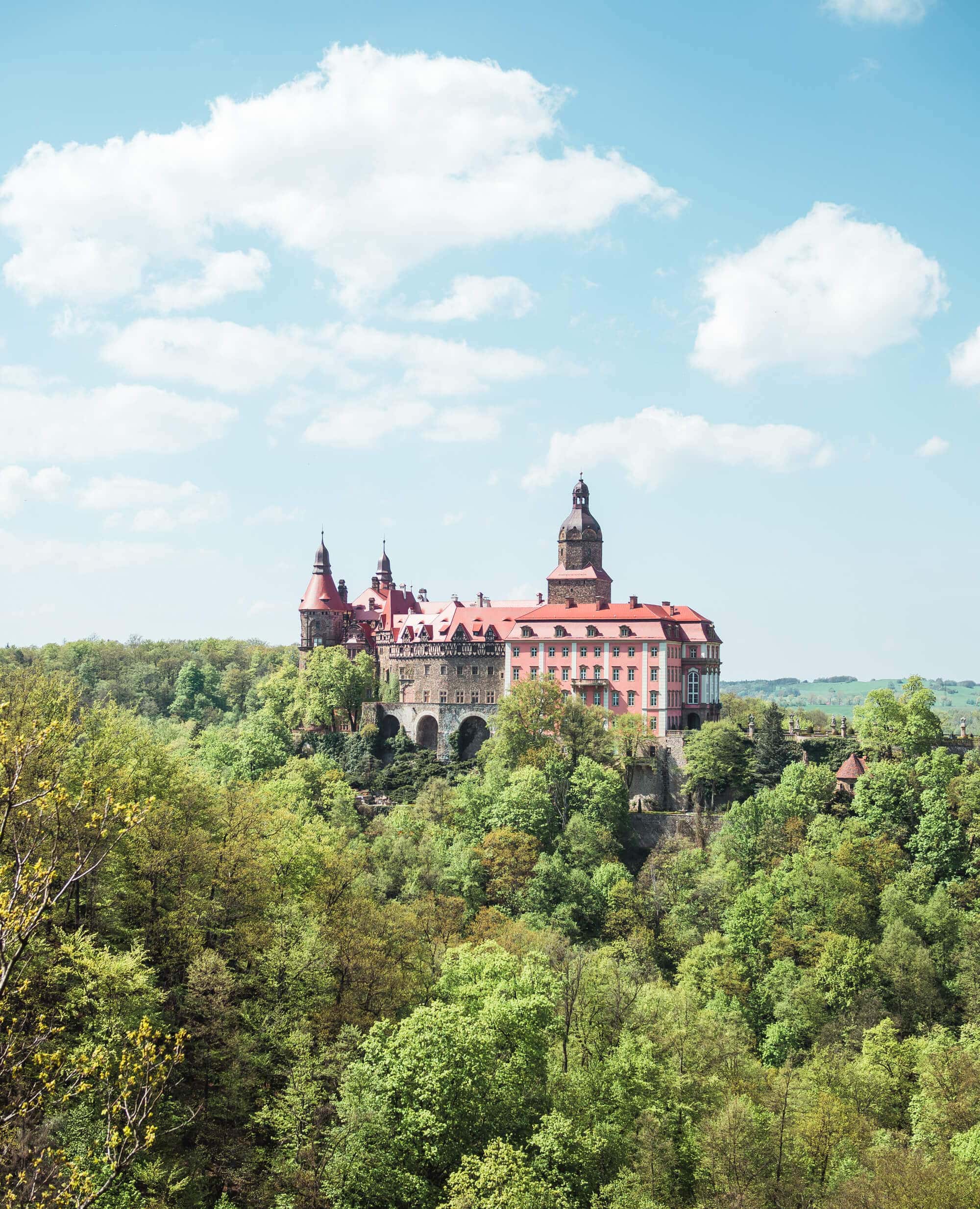 Pink fairytale-esque Książ Castle set on a hill amongst lush greenery, one of the most beautiful castles in Poland.