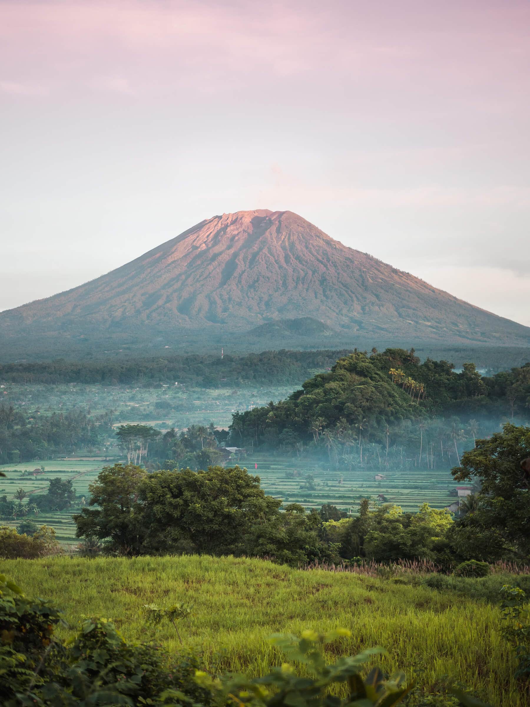 View of Mount Agung volcano seen from Bukit Cinta viewpoint in East Bali at sunrise against a pink sky and lush green rice fields in the foreground.