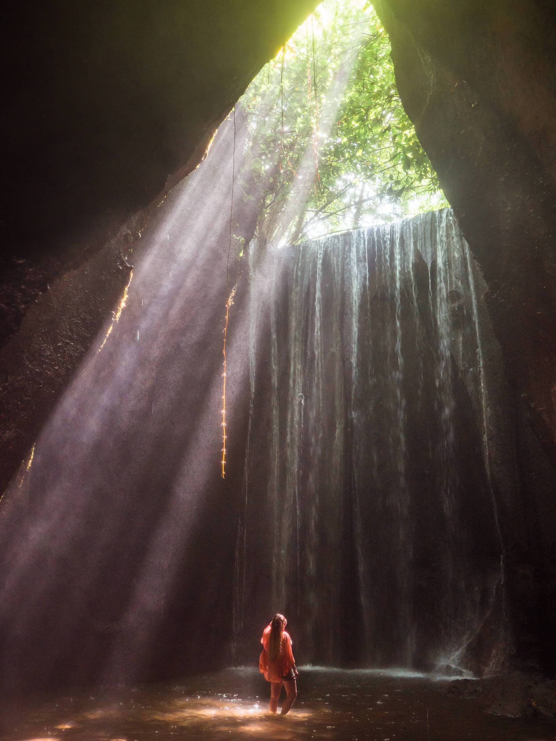 Woman wearing a red kimono standing in the pool at the bottom of Tukad Cepung waterfall inside a cave with the sun shining in from the top, one of the top things to do in Bali.