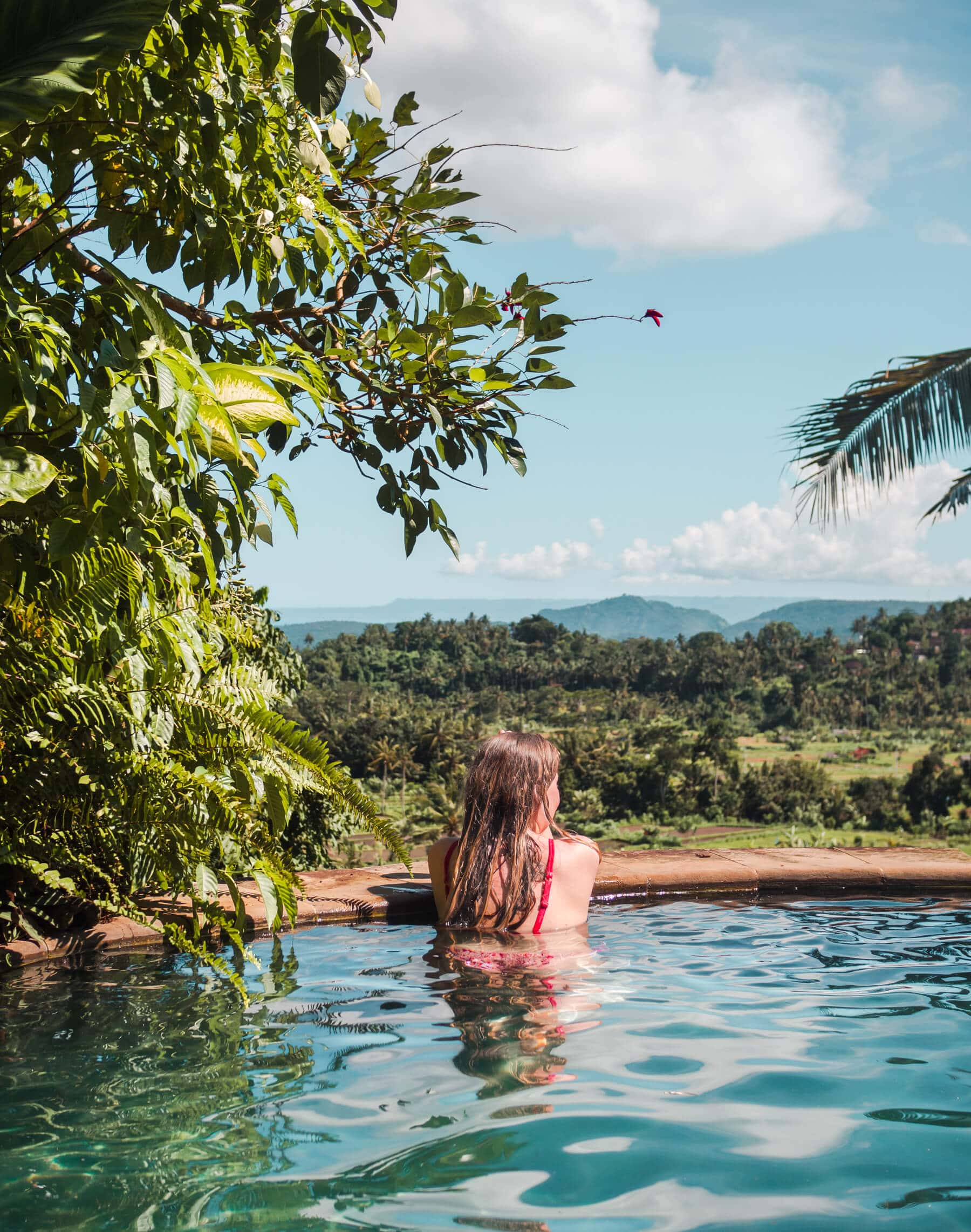 Girl with long hear wearing a red bikini lokking over the edge at the pool at Kubu Carik Bungalows, over the lush jungle and rice fields.