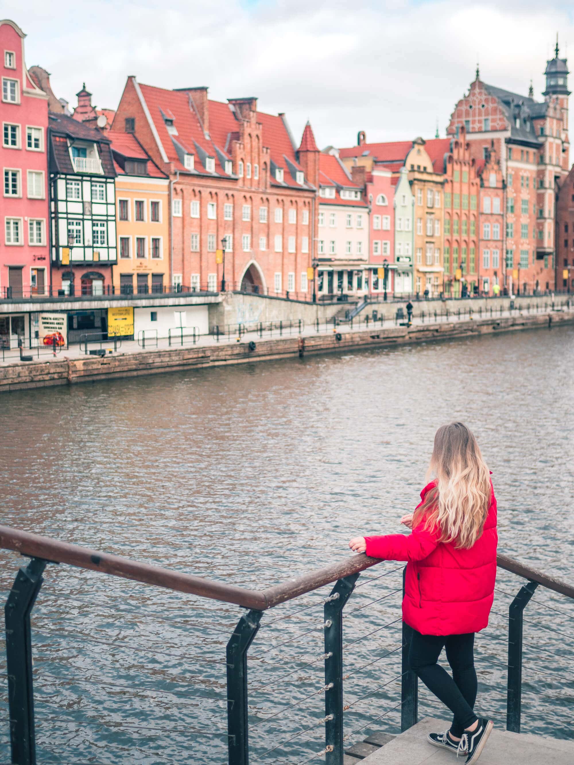 Girl with long hair, wearing a red down jacket and black pants, looking out over Gdansk River, with a row of colorful houses on the other side, a must during your 2 days in Gdansk.