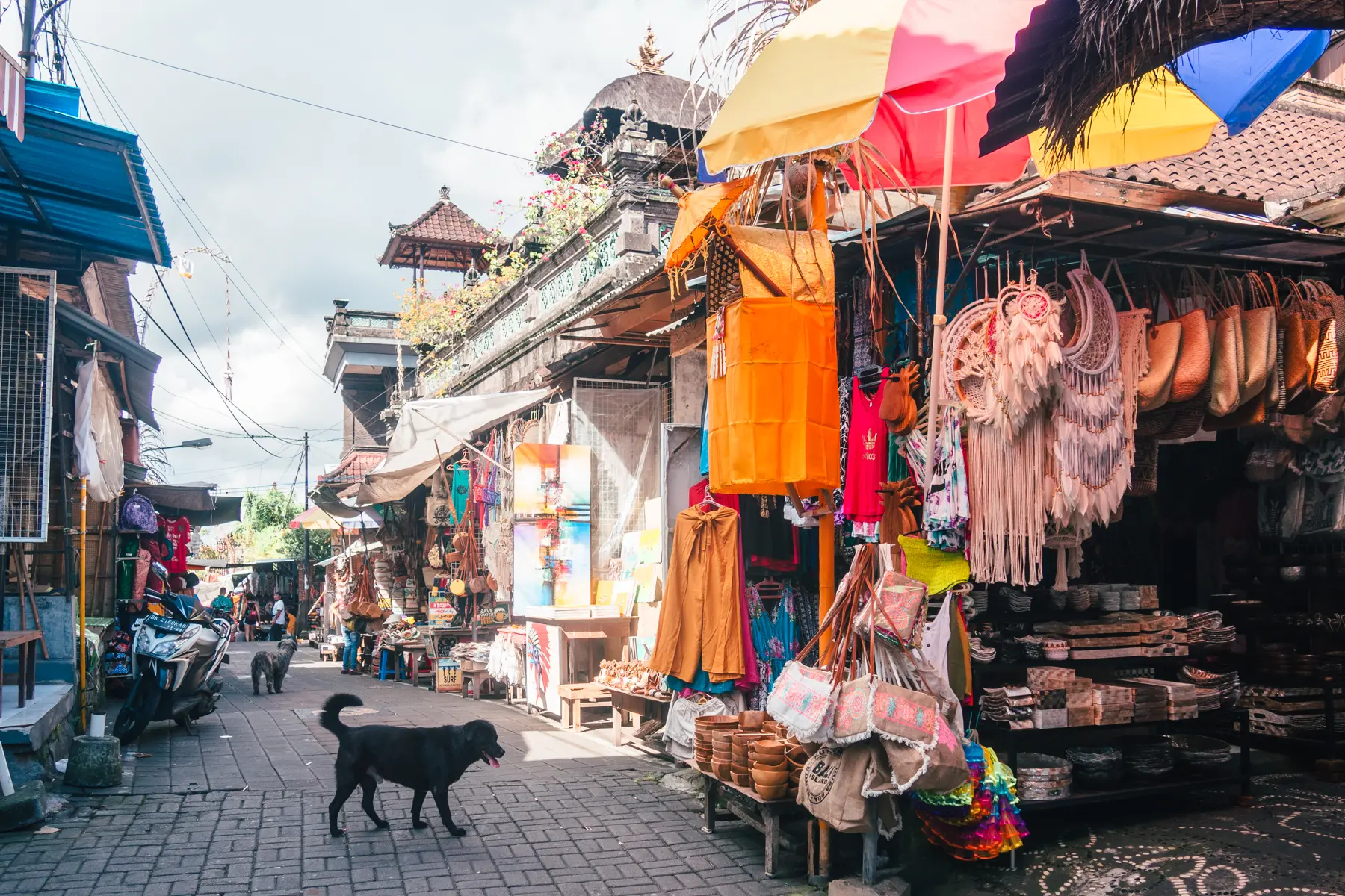Black dog walking across an alley at Ubud Art Market with stalls selling souvenirs at both sides.