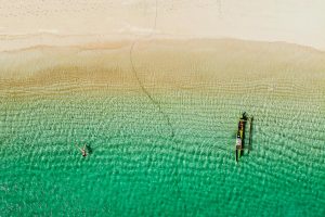 Drone shot of a girl and boat floating in green water at Tanjung Aan Beach, one of the best beaches in Kuta Lombok.