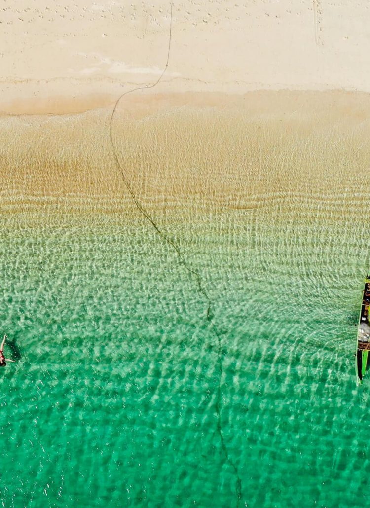 Drone shot of a girl and boat floating in green water at Tanjung Aan Beach, one of the best beaches in Kuta Lombok.