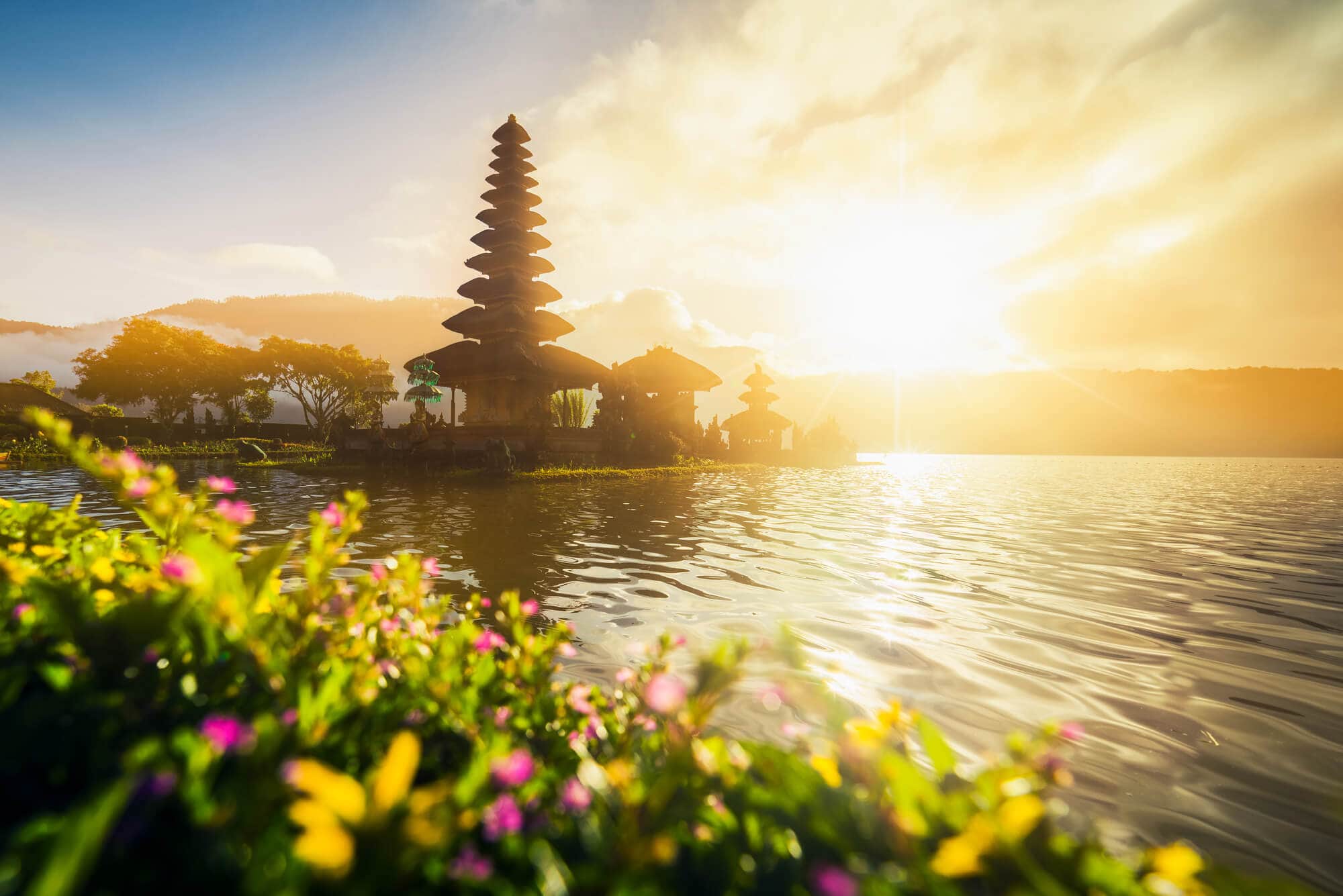 Glowing yellow sunset over Ulun Danu Beratan Temple in the middle of a lake with pink flowers in the foreground, a must on any 2-week Bali itinerary.