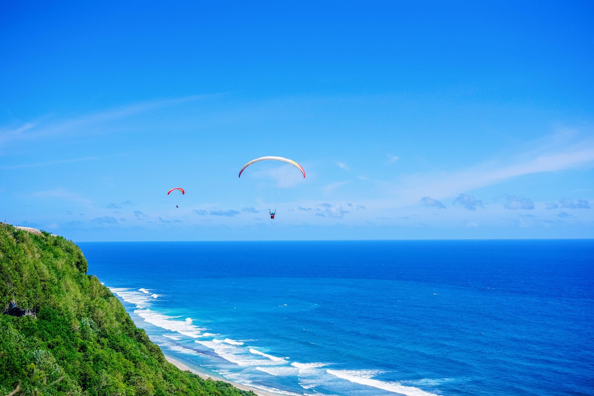 Two paragliders against a clear blue sky, above the blue ocean off Bali's Nyang Nyang Beach with lush green cliffs to the left. Paragliding is now included in SafetyWing Nomad Insurance.