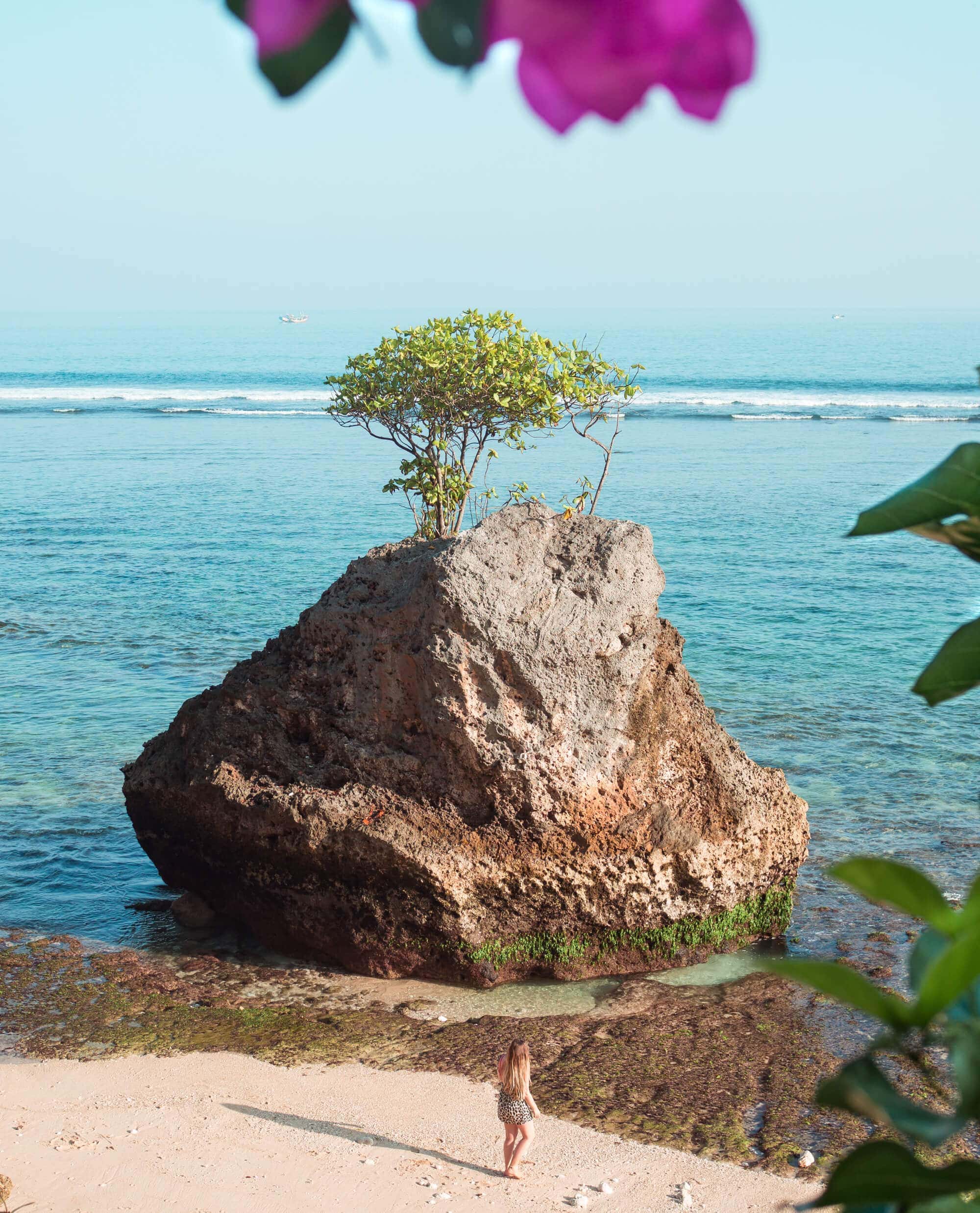 Girl standing on the water's edge on Bingin Beach at sunset with a large rock in front of her. Small waves roll in the background, and the blue water and sky looks like one. Bingin Beach is a must on any two-week Bali itinerary.