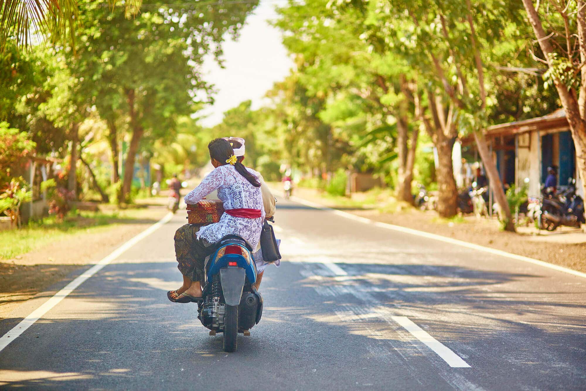 Man and woman wearing traditional Balinese clothing driving on a scooter on an empty road with greenery on both sides. How to get around during your 2-week Bali itinerary 