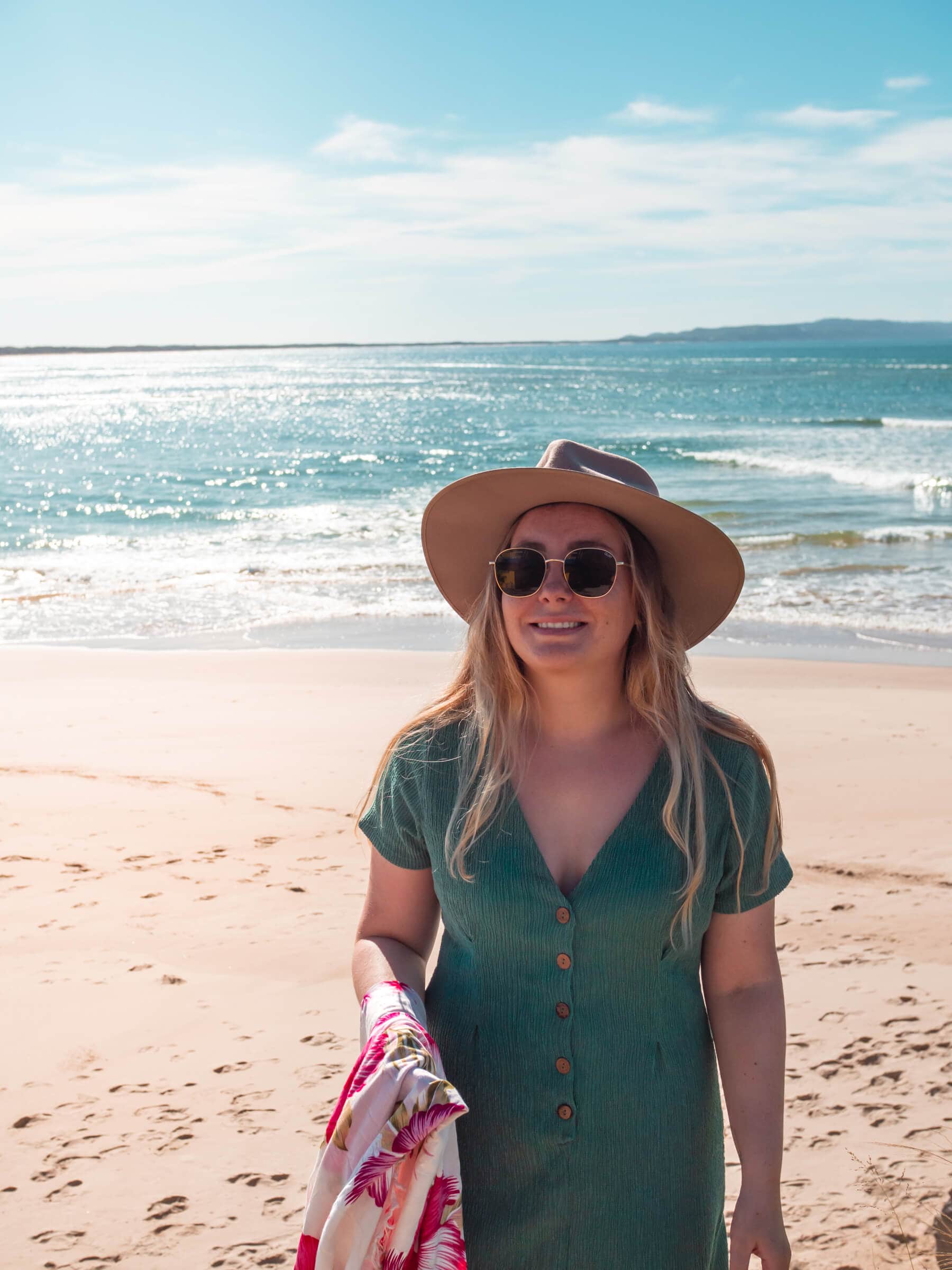 Girl with long dark blonde hair wearing a beige hat, sunglasses and a green dress smiling on the beach in Noosa.