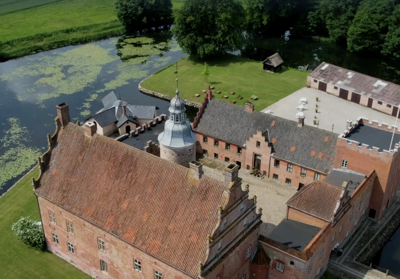 Aerial view of red brick castle compound next to green lawns and a pond, Broholm Castel Hotel in Denmark.