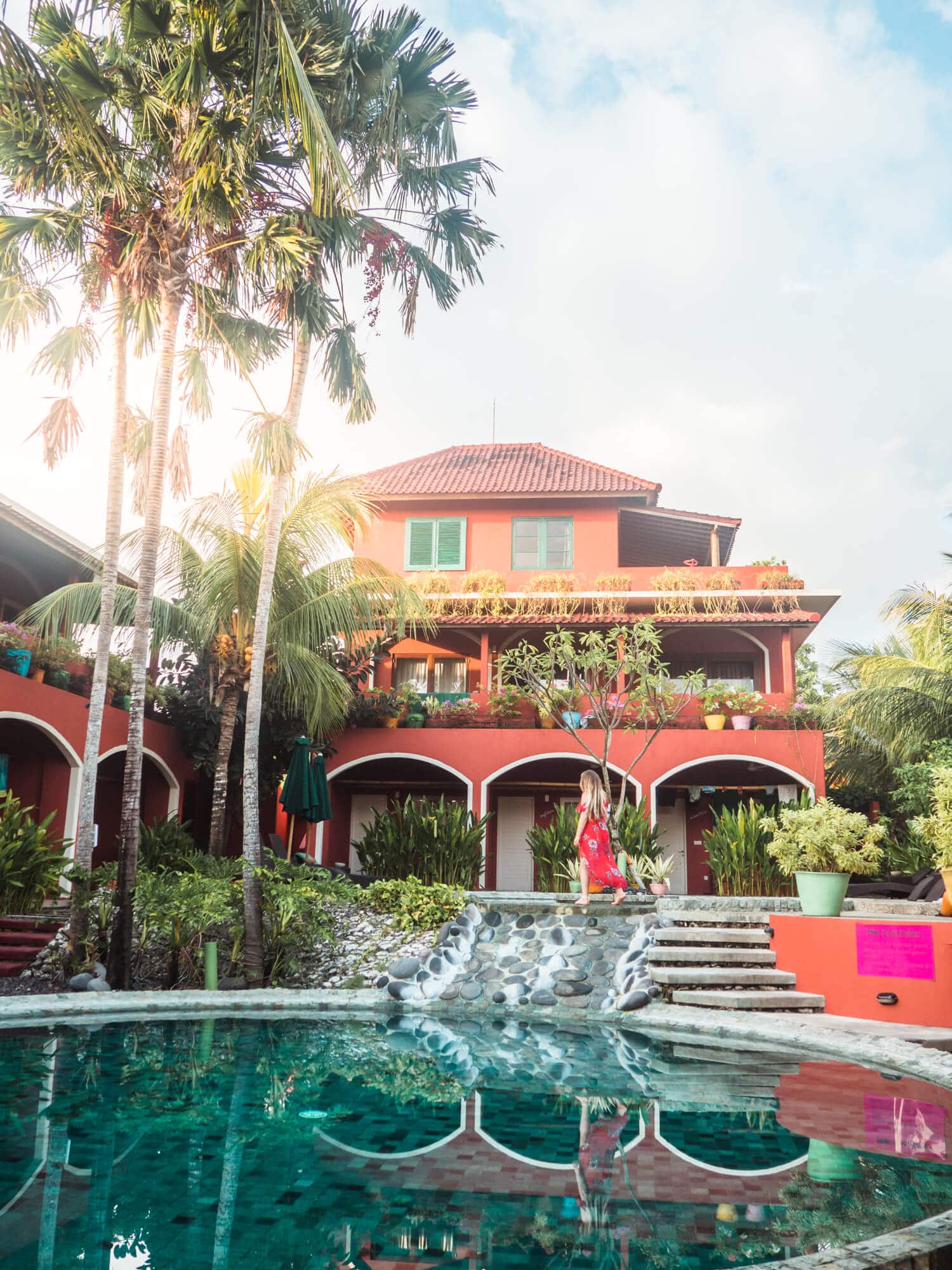 Girl in a red dress walking in front of Pink Coco Hotel, with a green pool in the foreground and a three tall palm trees to the left. The best place to stay in Uluwatu during your two weeks in Bali.