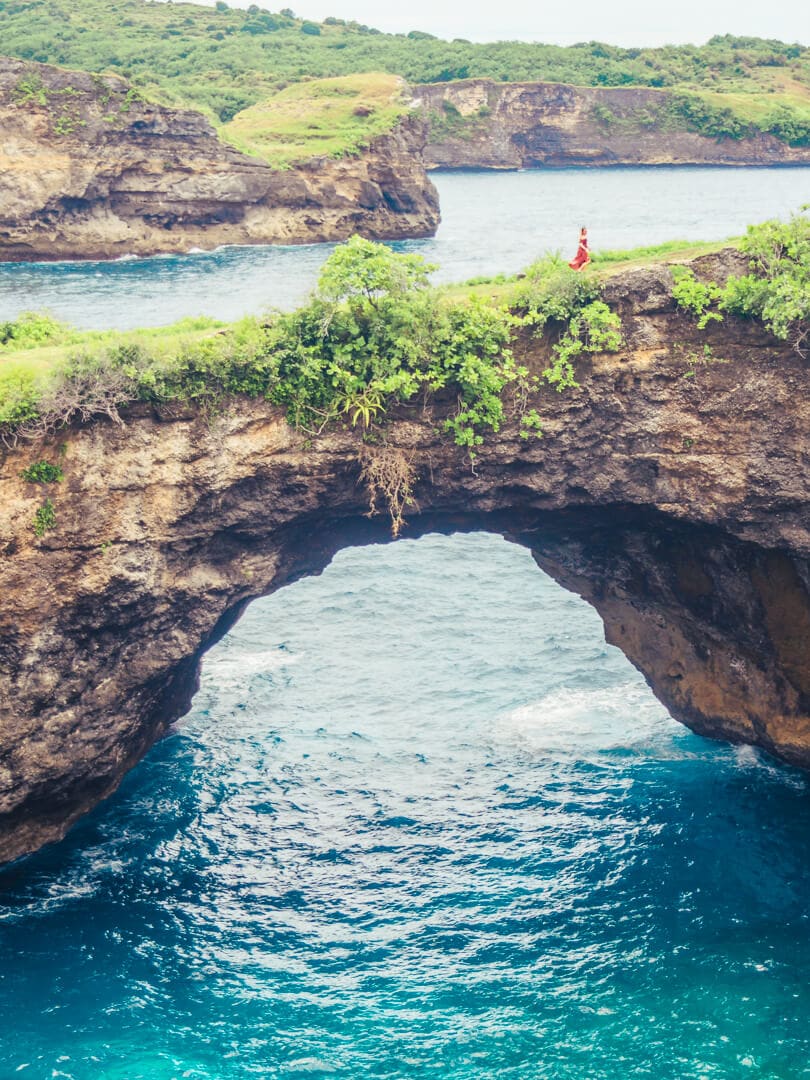 View of Broken Bean on Nusa Penida, a rock arch with greenery on top and blue water underneath. Girl in a red dress walking on top, on a day trip to Nusa Penida during her two week Bali itinerary.