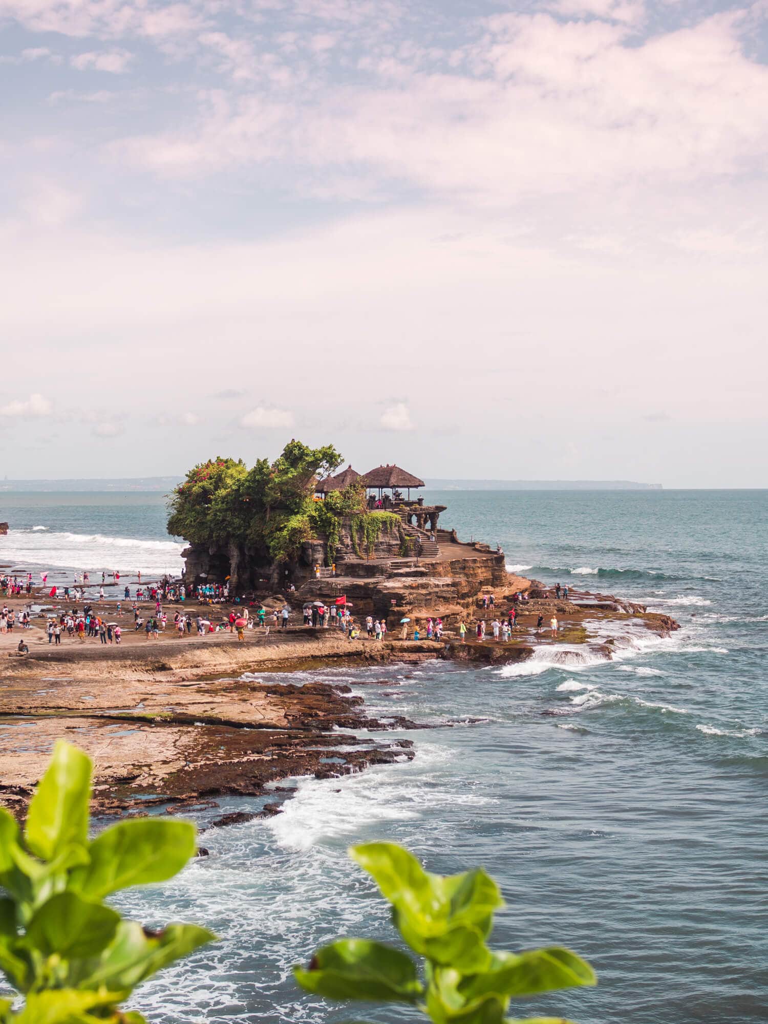 View of Tanah Lot temple set on a rocky outcrop in the water, just outside Canggu. The sky is turning purple for sunset as a large group of people make their way to the temple. A must on any 2-week Bali itinerary.