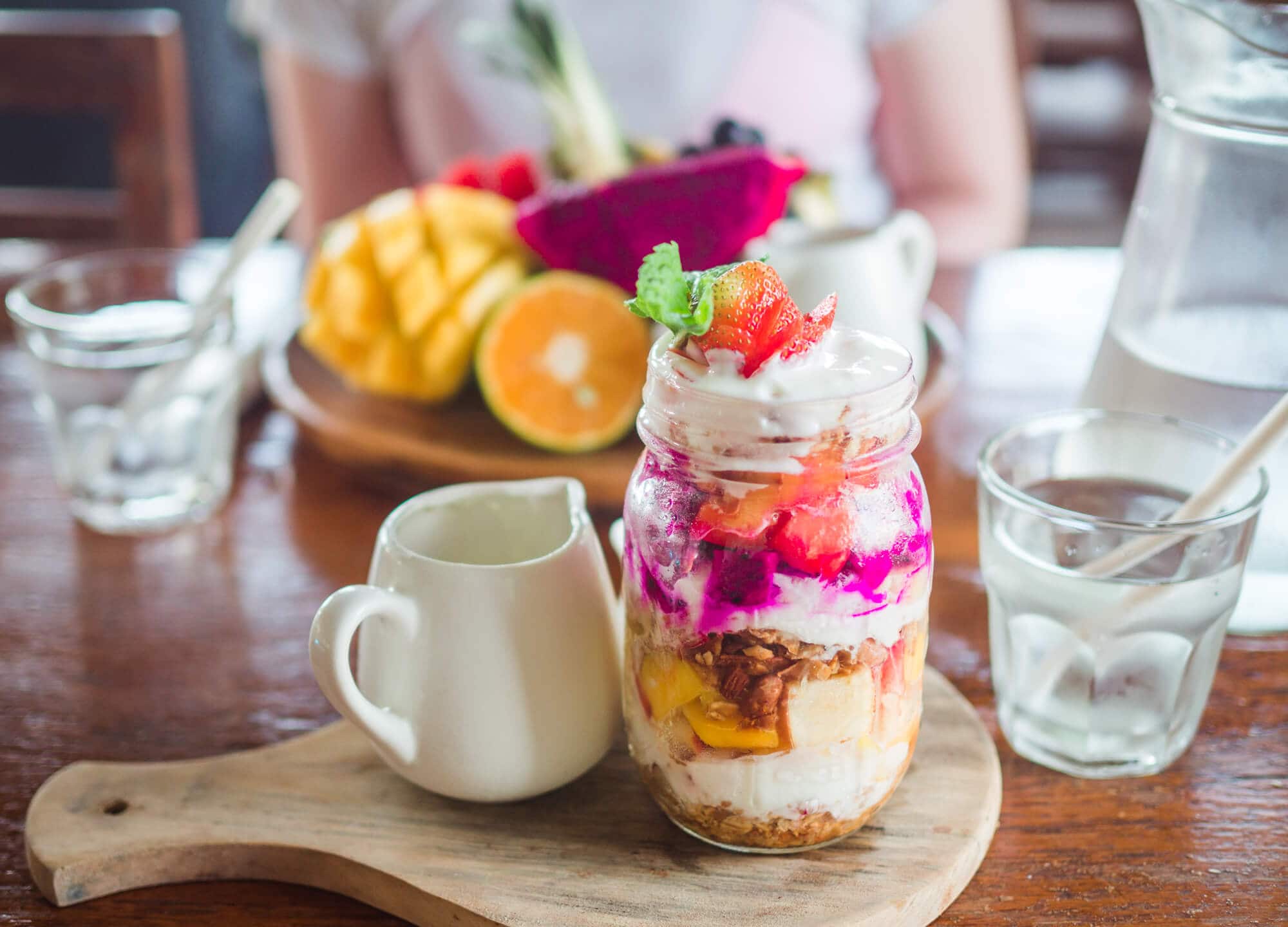 Parfait with pink dragonfruit and granola served in a glass with a large fruit platter in the background in Bukit Café, one of the best places to have breakfast in Uluwatu during your two weeks in Bali.