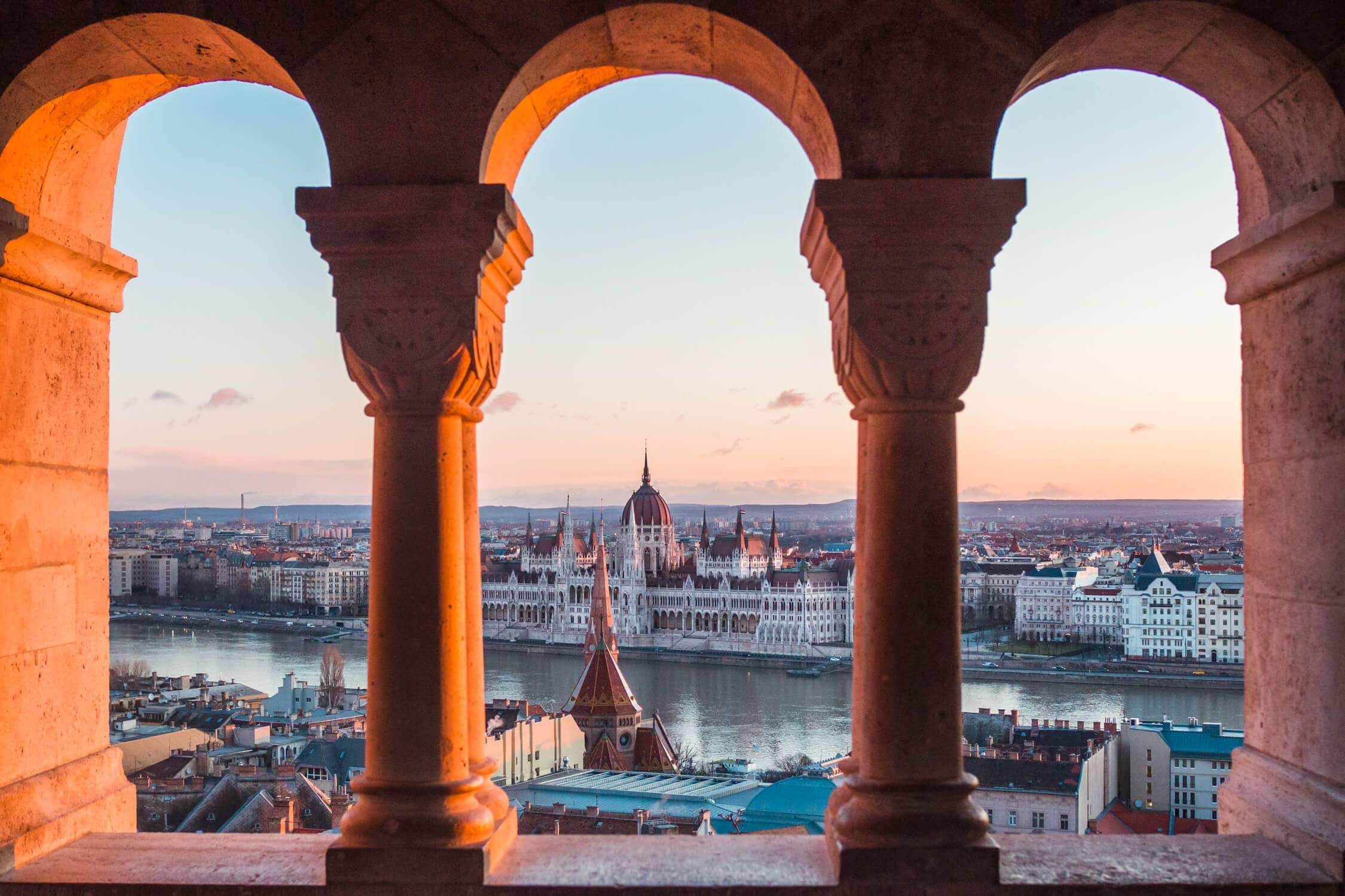 Early morning view of the Budapest Parliament building from Fisherman's Bastion, framed by three arches. Best Instagram spot in Budapest.