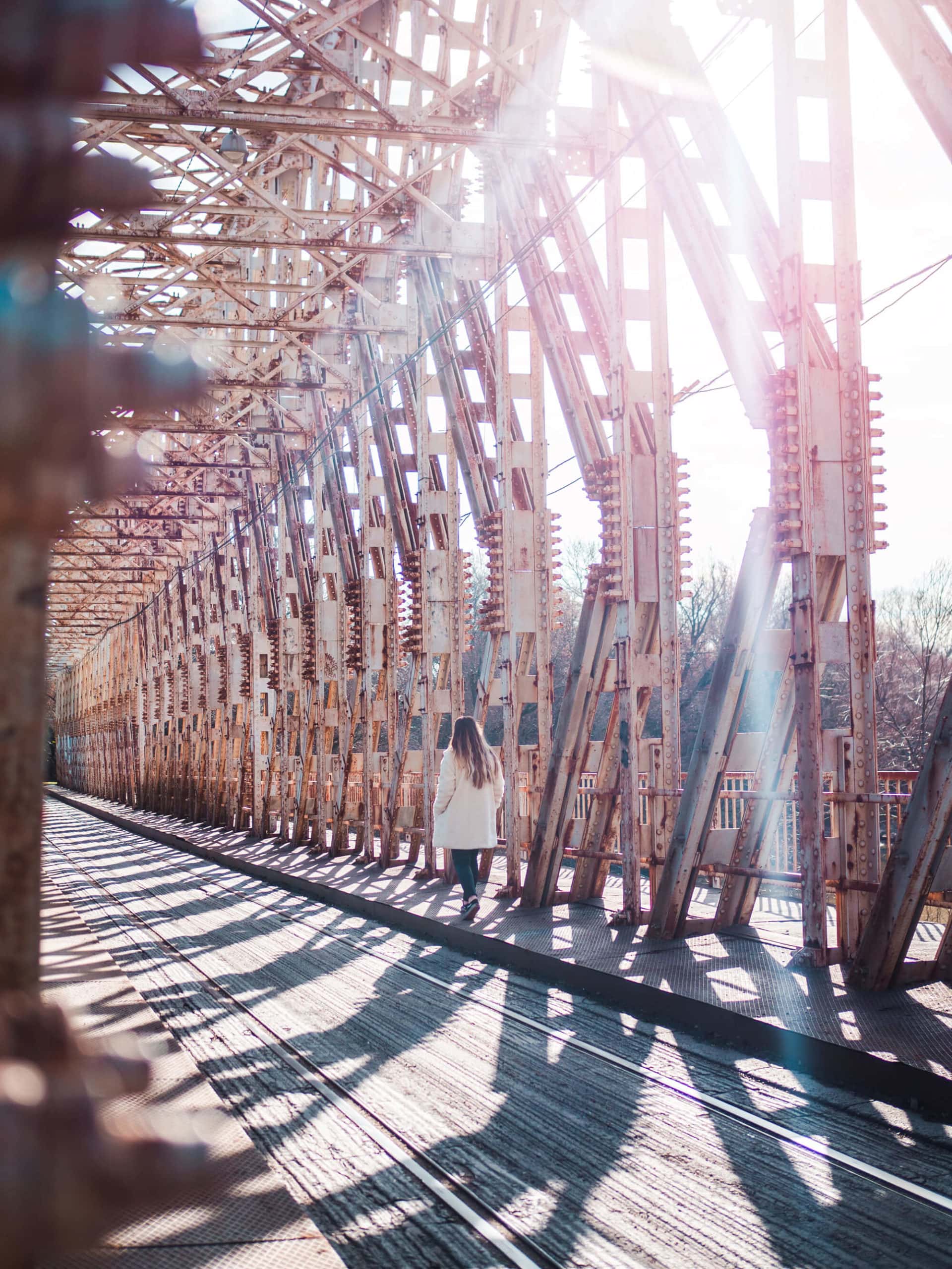 Woman wearing a white coat walking along a metal bridge with the sun shining in, using the Budapest Card. Was it worth it?