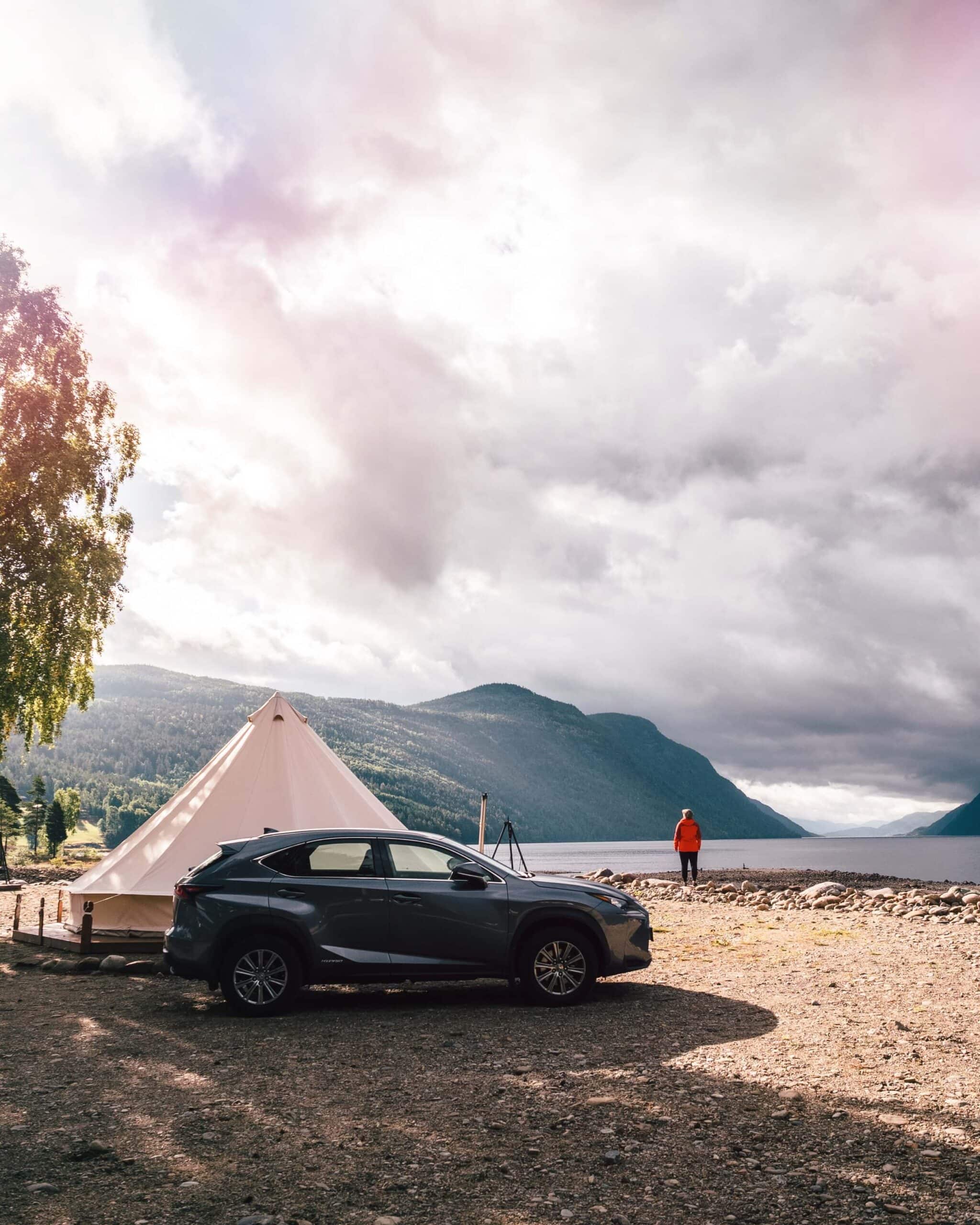 Grey Lexus outside a glamping tent at Sandviken Campin in Norway on a cloudy day with a woman in a red jacket standing by the waters edge, to cut down on the travel cost in Norway.