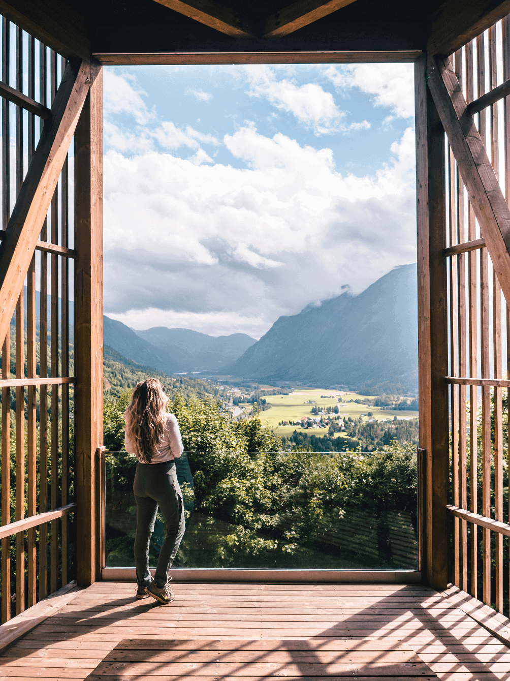 Girl with long hair, wearing green hiking pants and a white top, standing at Flatdal Viewpoint in Norway, looking out over a lush green valley with mountains on both sides. The cost of travel in Norway.