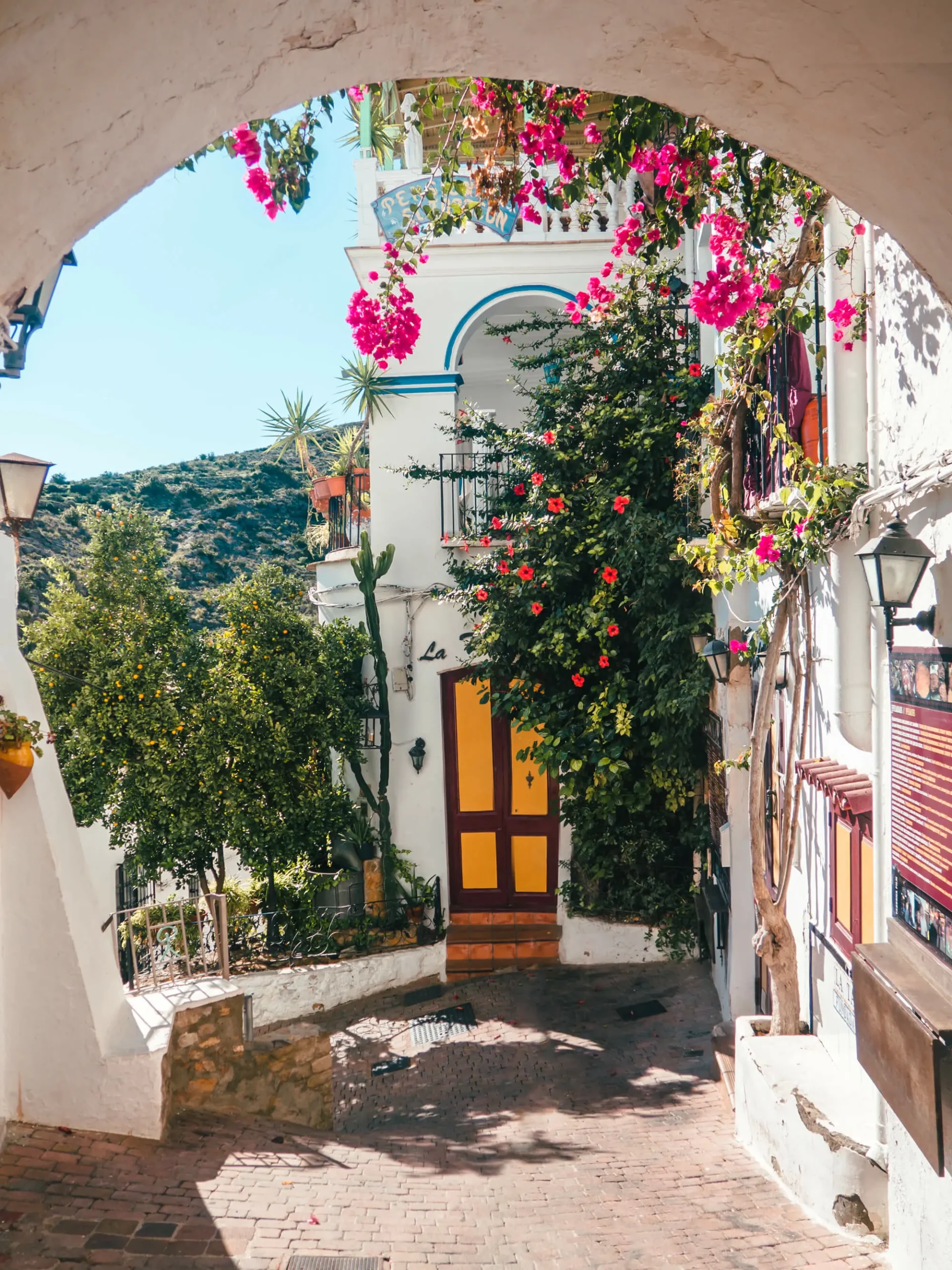 Looking at a red and yellow door on a white house covered in pink Bougainvillea through a white arch in Mojacar Pueblo, one of the most beautiful white villages in Spain.