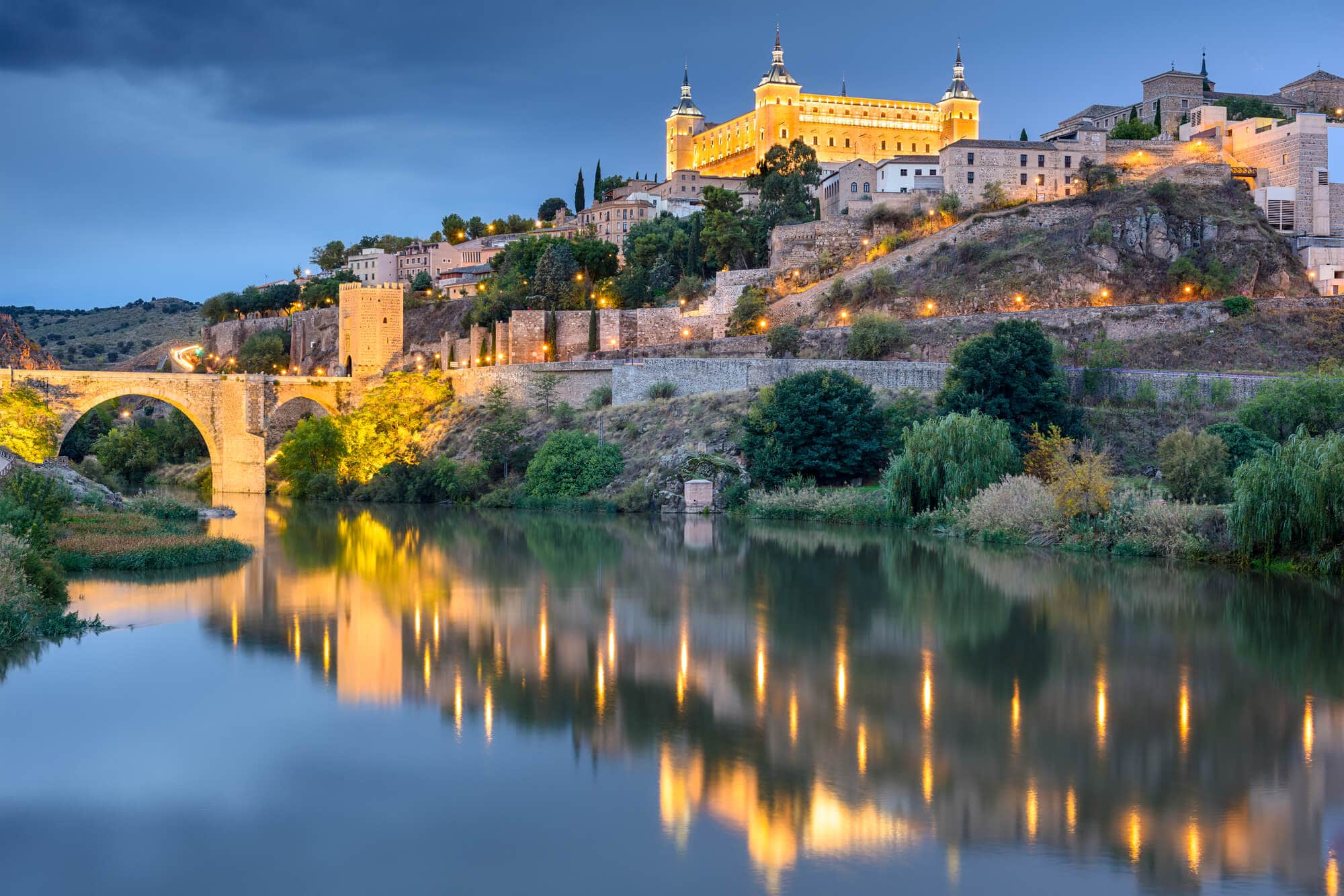 The beautiful Alcázar de Toledo lit up at dusk seen from the banks of the Tagus River - The Ultimate Spain Bucket List