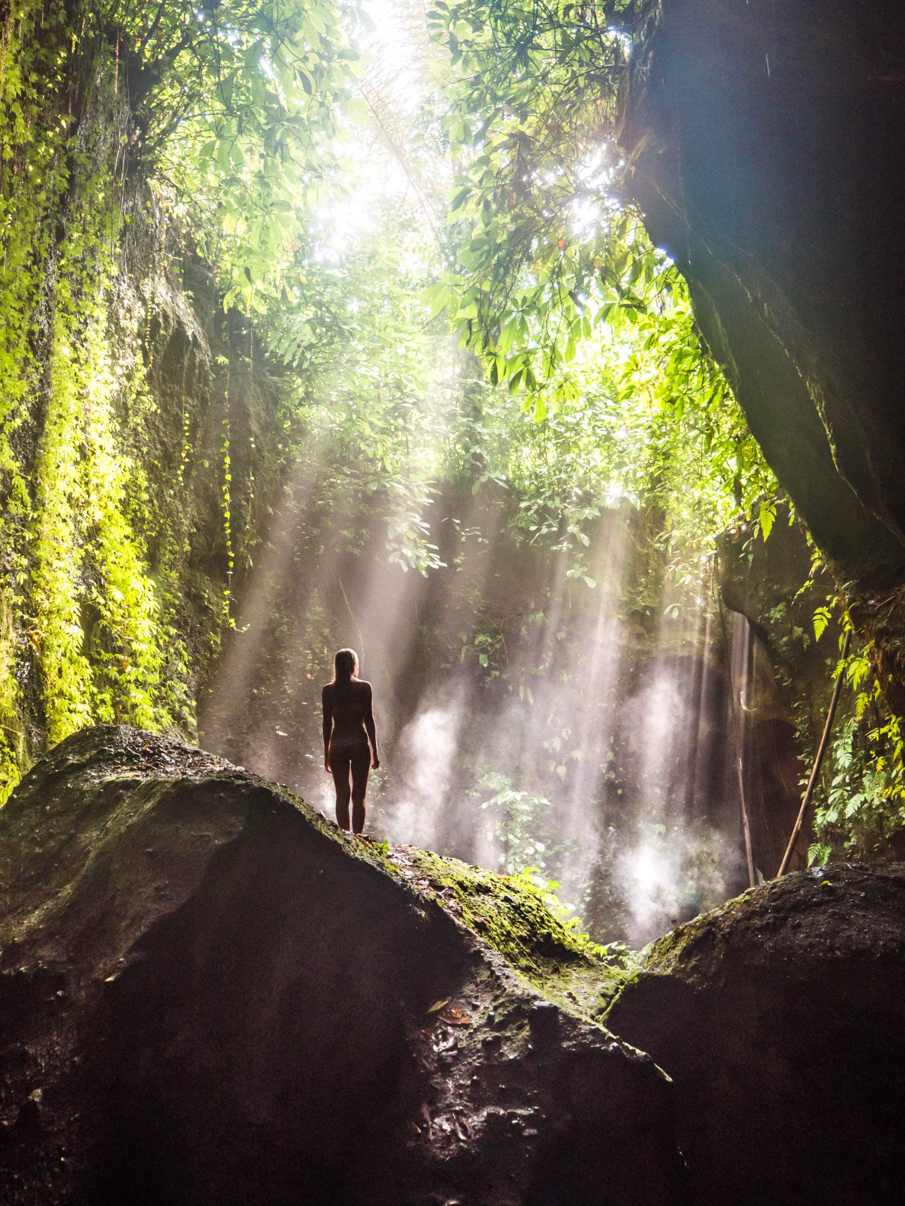 Girl stanidng on a rock in Tukad Cepung Waterfall with sun flares coming through the greenery in Ubud Bali