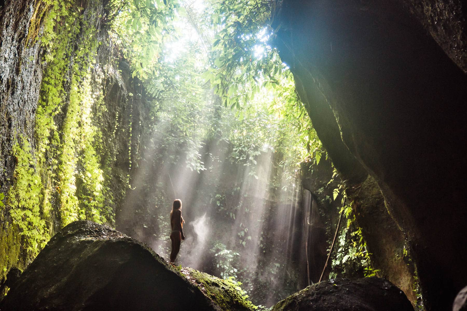 A guide to all the best waterfalls in Ubud Bali - A girl standing on a rock at Tukad Cepung Waterfall with the sun shining into the cave