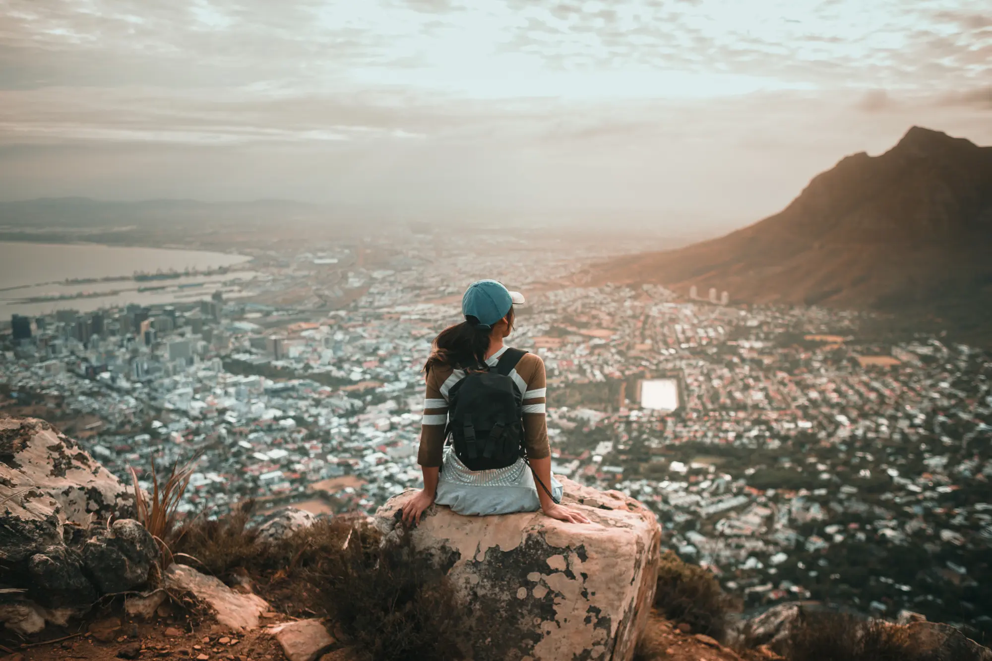 Woman with black hair wearing a baseball cap, hiking gear and black backpack, sitting on the edge of a cliff looking out over a city below a mountain.