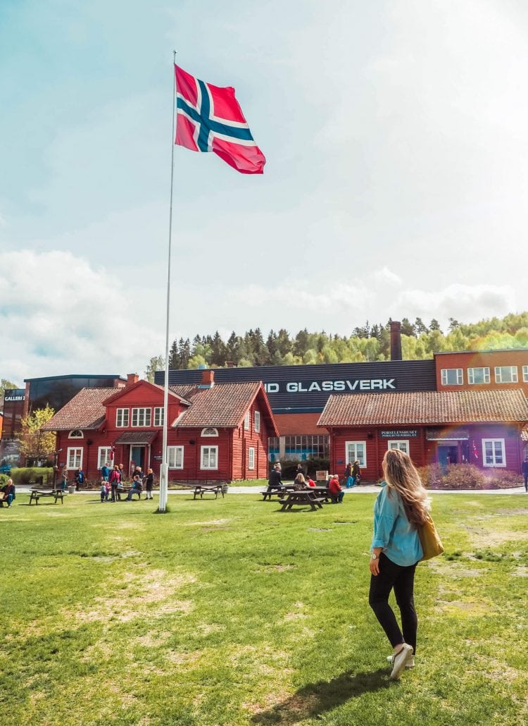 Woman with long hair, wearing a denim shirt and black pants, in front of a white flagpole with the Norwegian flag on a green lawn, red and white old traditional buildings at Hadeland Glassverk, the perfect day trip from Oslo Norway.