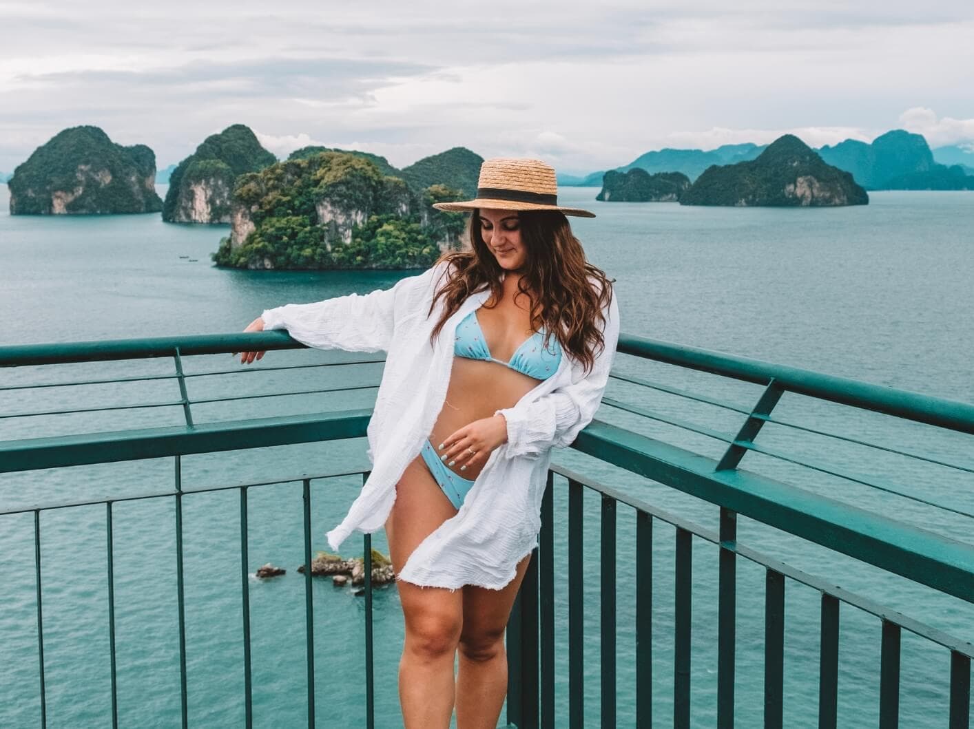 Girl with long dark hair wearing a beige hat, blue bikini and white shirt standing at a viewpoint overlooking turquoise water and islets during a tour to Hong Island from Krabi.