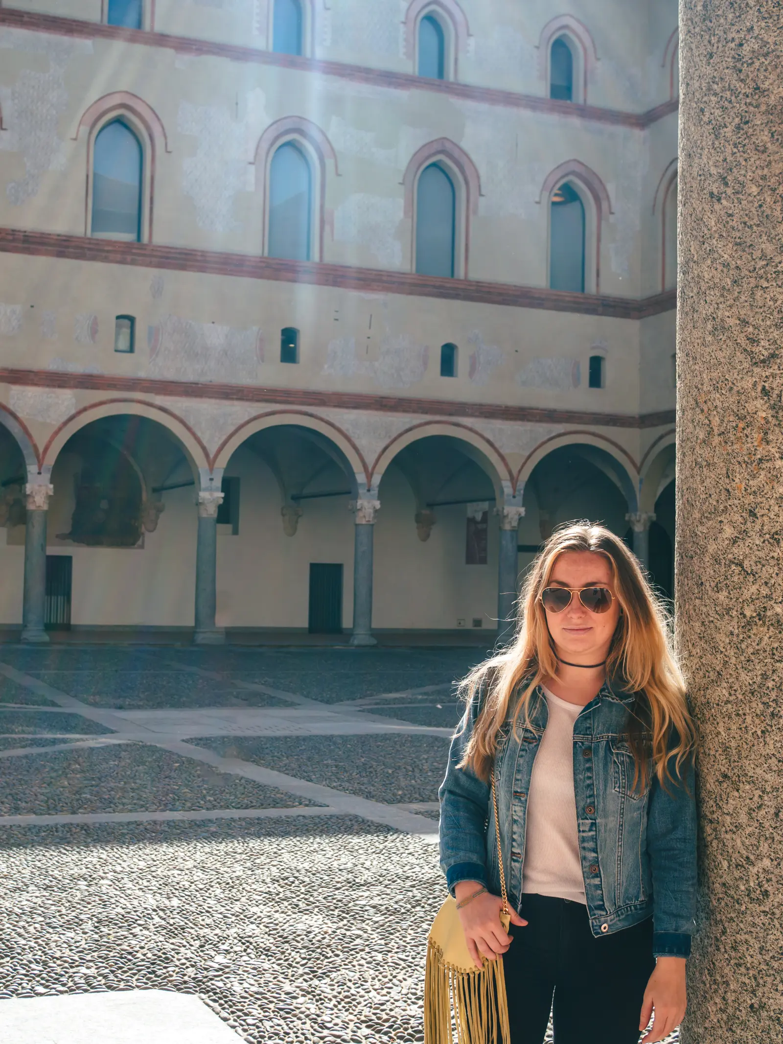 Girl wearing a denim jacket, white t-shirt, black pants and yellow bag standing against a wall with and old building of arches during 2 days in Milan Italy.
