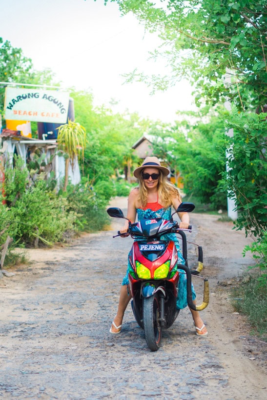 Woman with long hair, wearing a beige hat, red bikini top and blue sarong, driving a red scooter with yellow lights on a bumpy road on Nusa Lembongan surrounded by greenery.