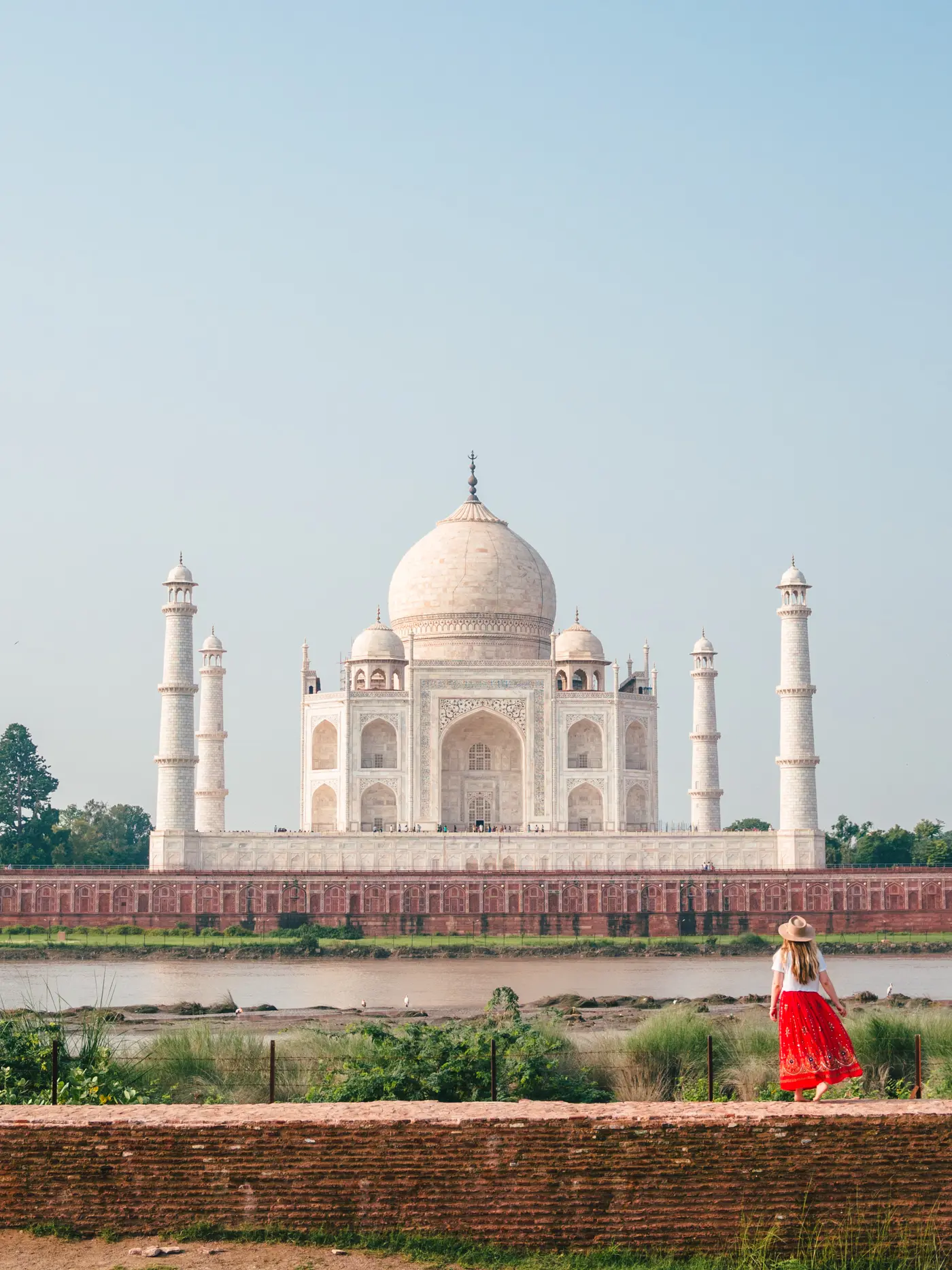 Girl wearing a red skirt and hat walking on a brick wall in Mehtab Bagh with Taj Mahal in the background.